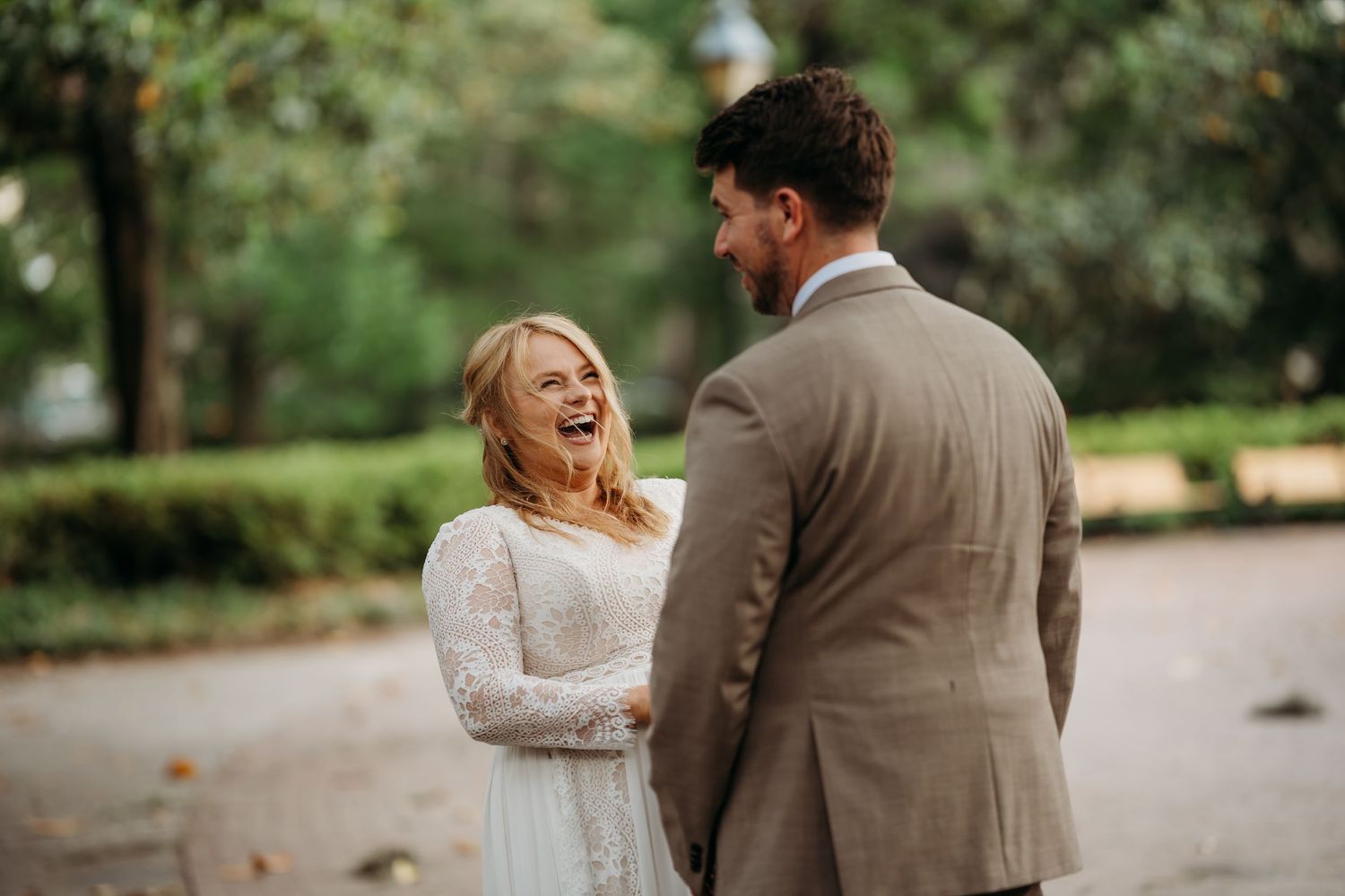 Joyful bride in lace wedding dress laughs with groom in tan suit during outdoor park celebration.