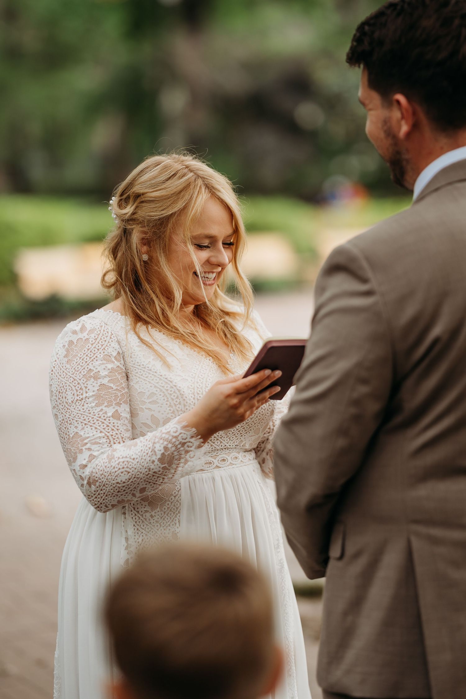 Smiling bride in lace gown reads wedding vows from a small book during outdoor park wedding ceremony.