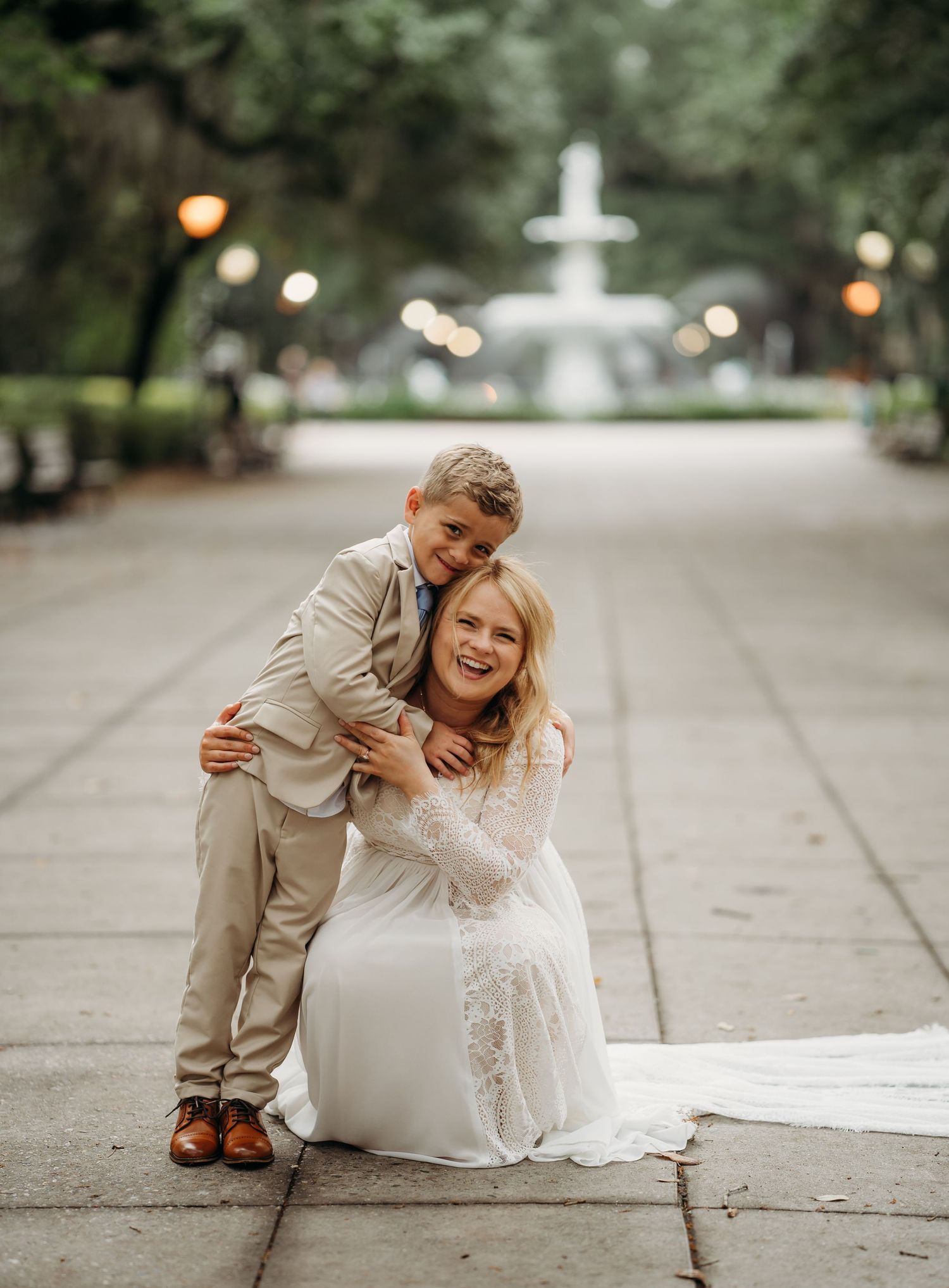 Laughing bride sits on Forsyth Park pathway as young boy hugs her from behind near fountain.