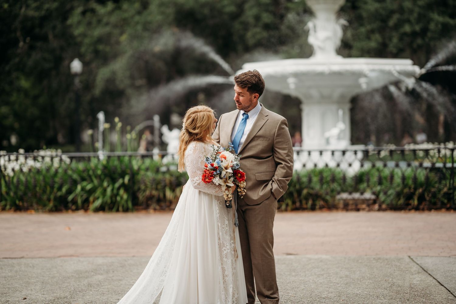 Bride and groom gaze at each other romantically in front of a large white park fountain.