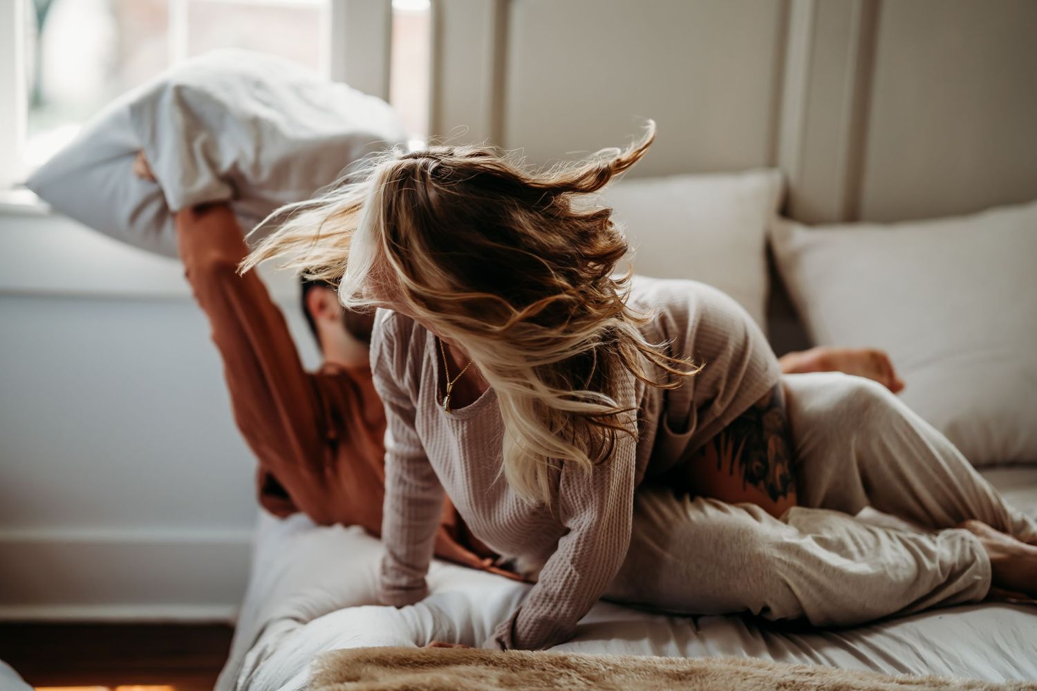 A woman with flowing blonde hair playfully tumbling on a cozy bed with her partner nearby.