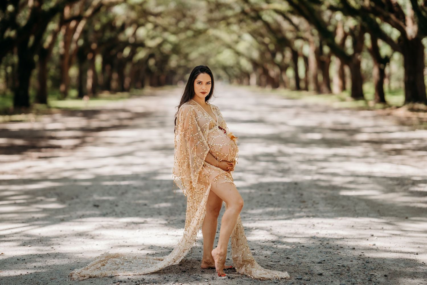 Pregnant woman in elegant gold lace maternity gown posing on a tree-lined path during an outdoor maternity photoshoot.