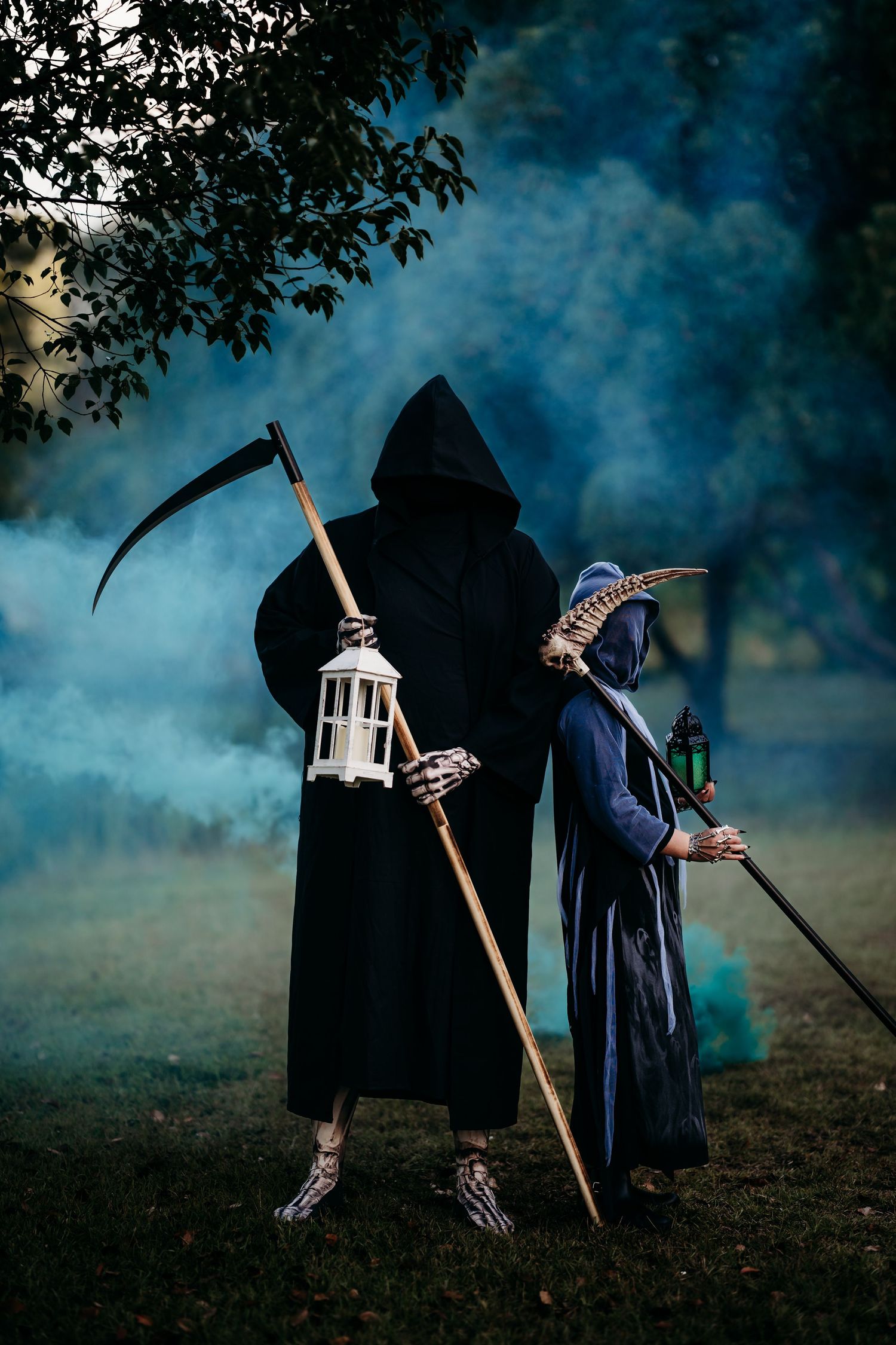 Two people in Halloween costumes as grim reaper and plague doctor holding lanterns in smoky forest.