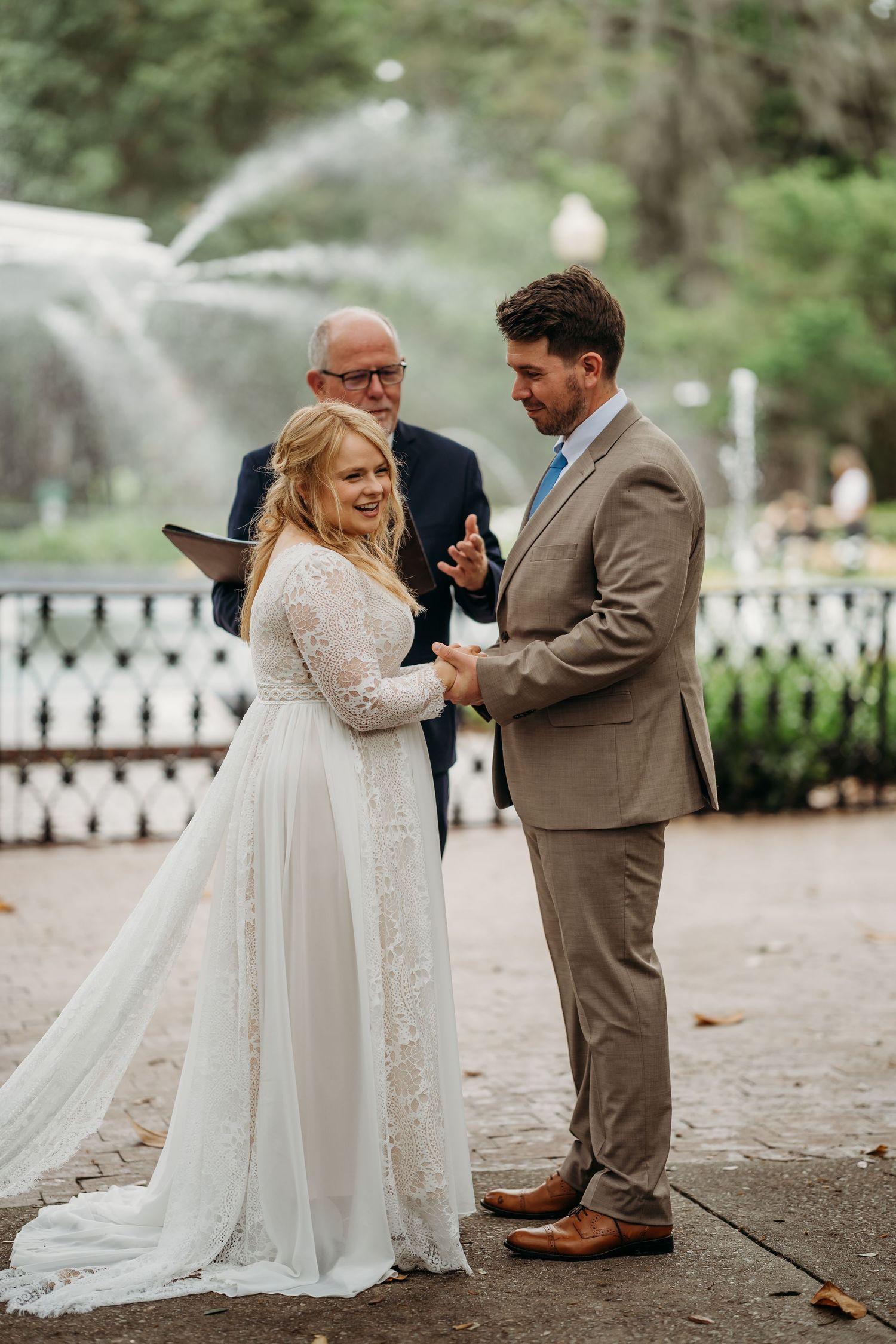 Smiling bride and groom hold hands as officiant speaks during outdoor wedding ceremony at Forsyth Park fountain.