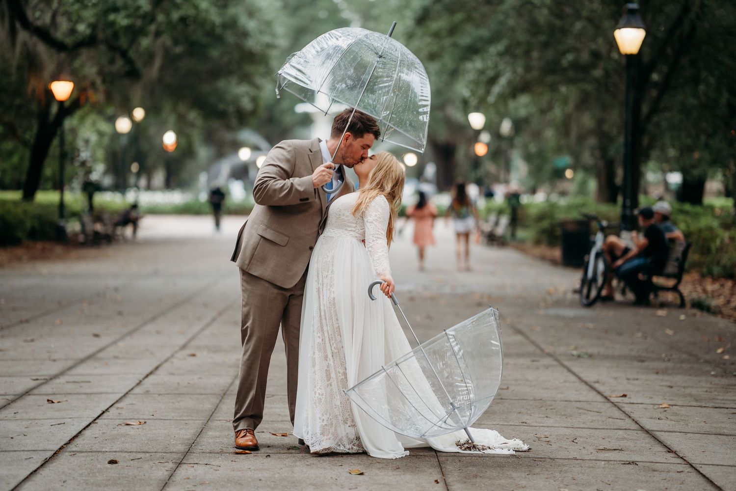 Wedding couple kissing under a transparent dome umbrella in a scenic tree-lined park pathway.
