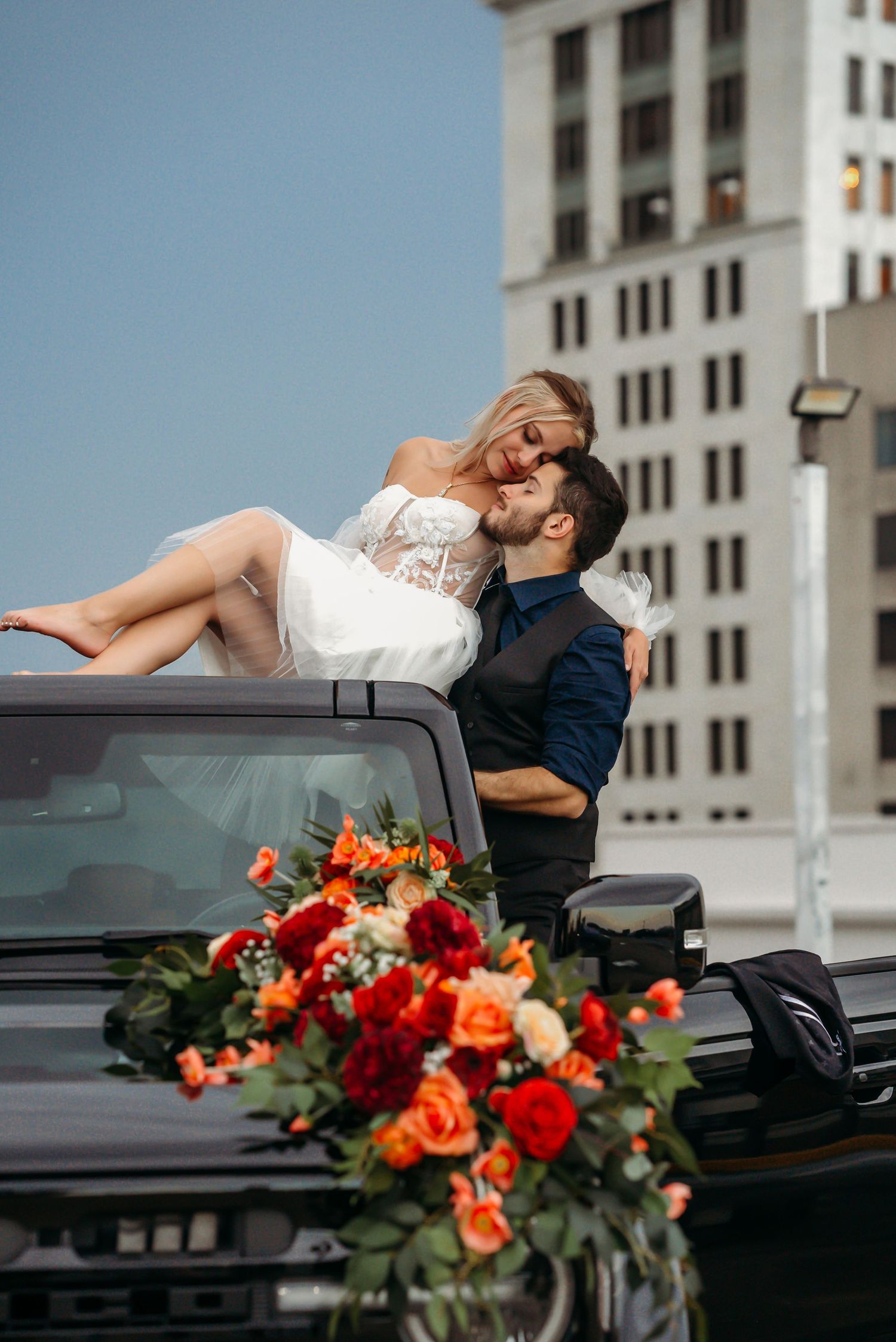 Bride in white dress held by groom on car rooftop with colorful floral arrangement in urban setting.