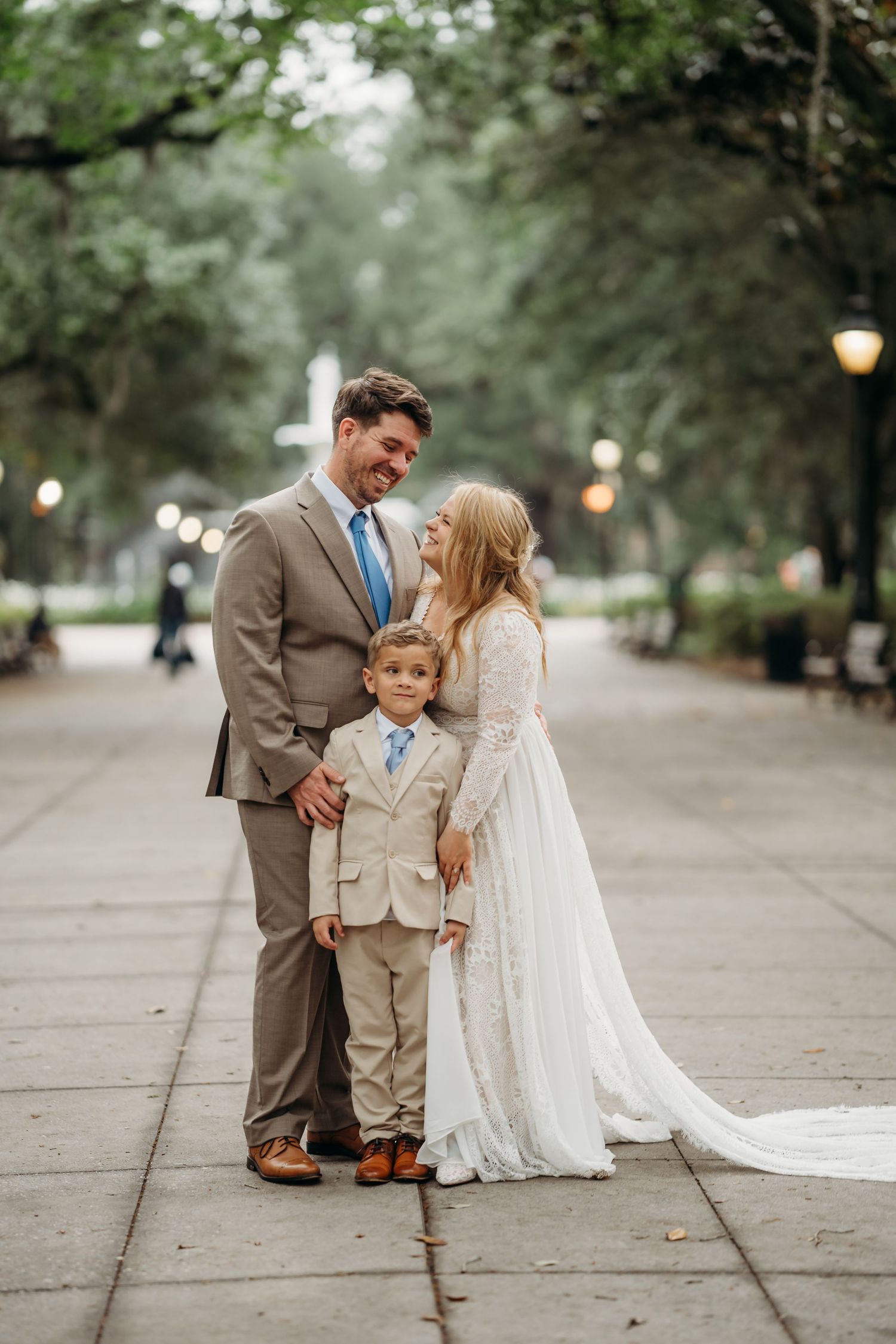 Bride and groom smile with young boy in matching tan suit at Forsyth Park, Savannah wedding.