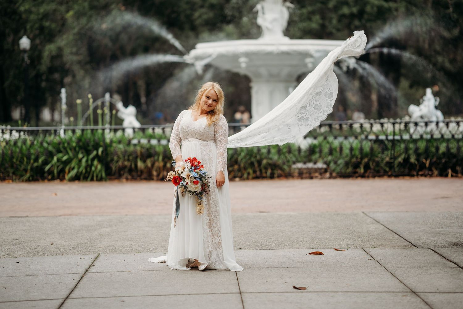 Bride in lace wedding gown with flowing veil caught in wind stands before ornate fountain holding bouquet.