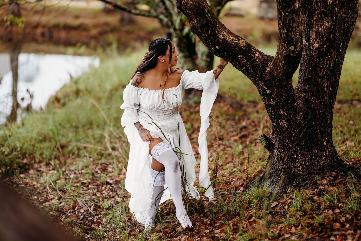 Woman in white off-shoulder gown and stockings leaning against a tree near a pond.
