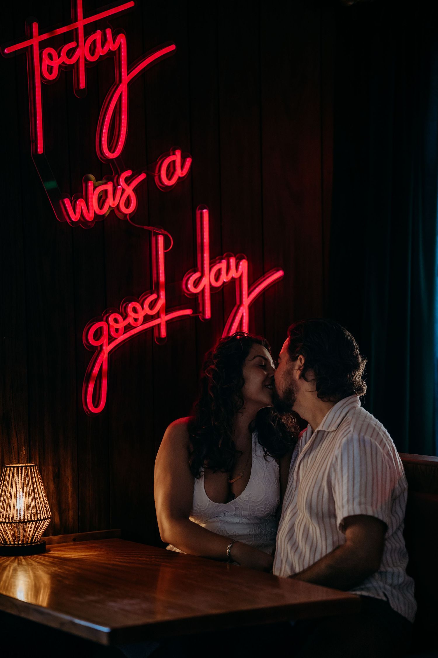 Couple kissing at table beneath red neon sign reading today was a good day.