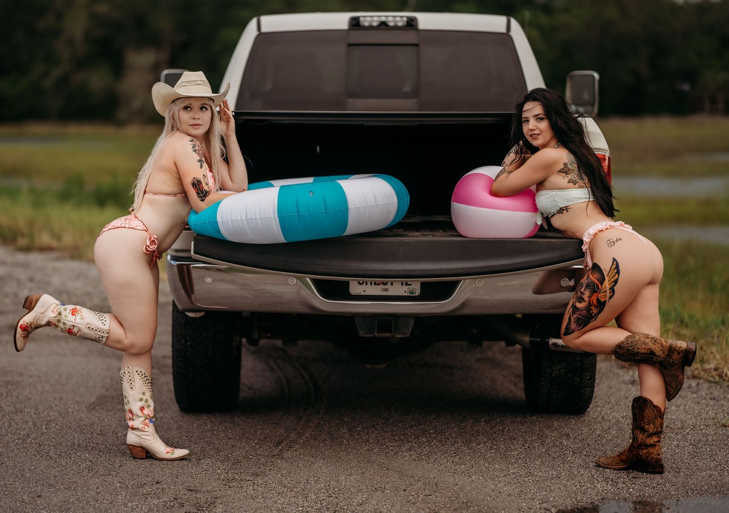 Two women in swimwear pose by a truck tailgate with colorful pool floats on a rural road.