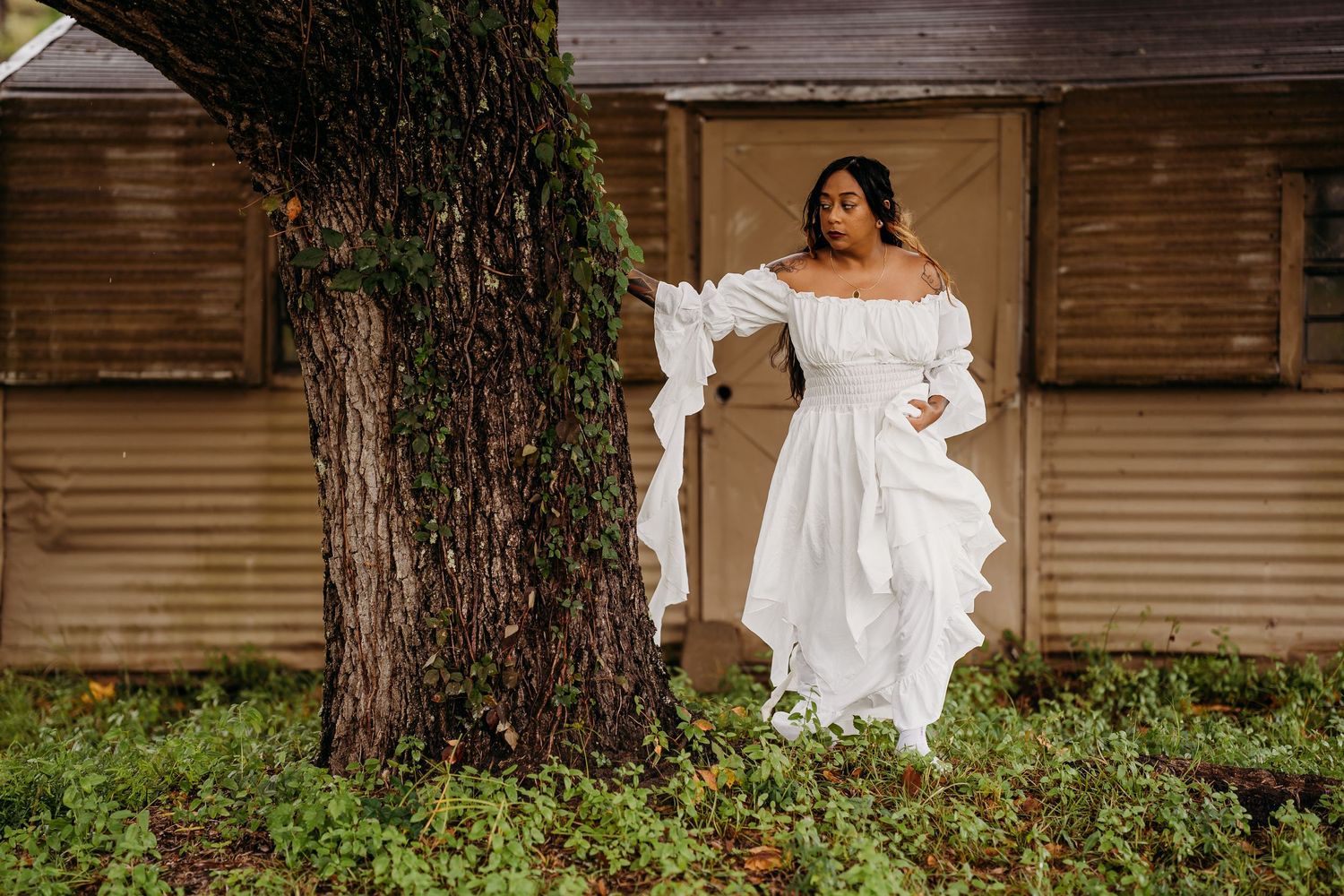 Woman in flowing white dress standing near a tree with fabric billowing by a rustic shed.