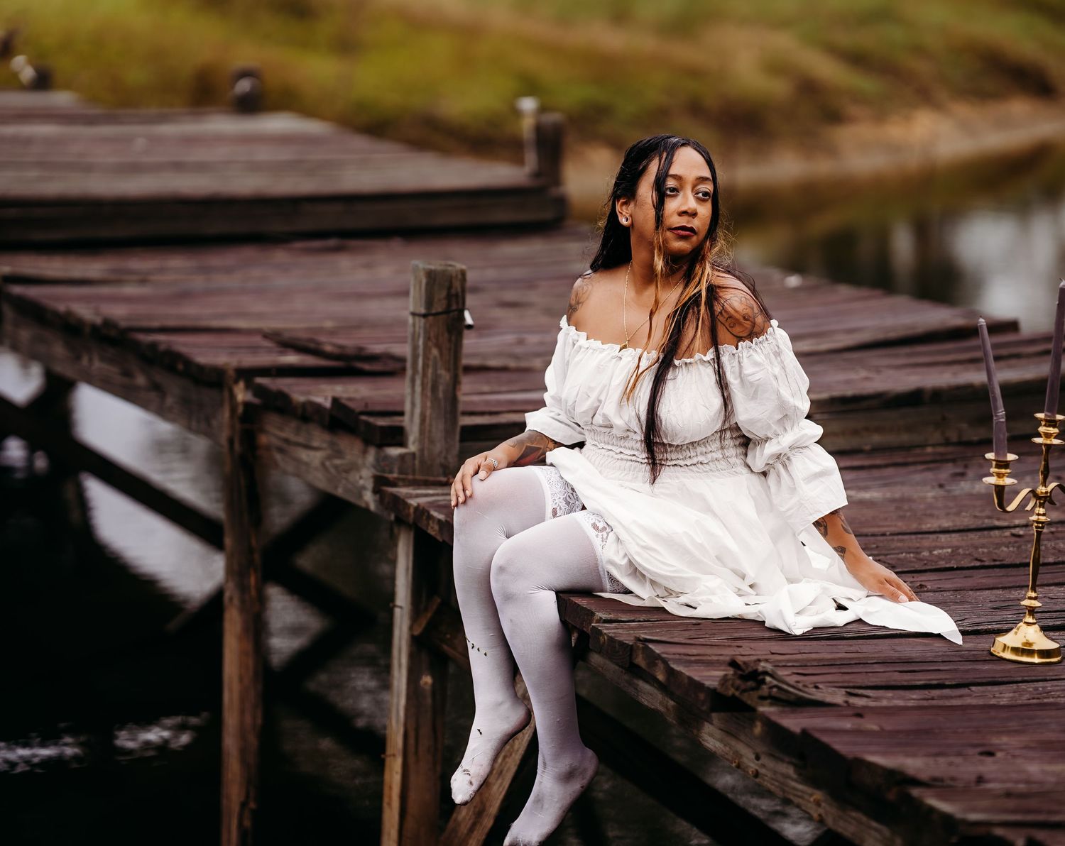 Woman in white off-shoulder dress and white stockings sits poised on wooden dock by water.