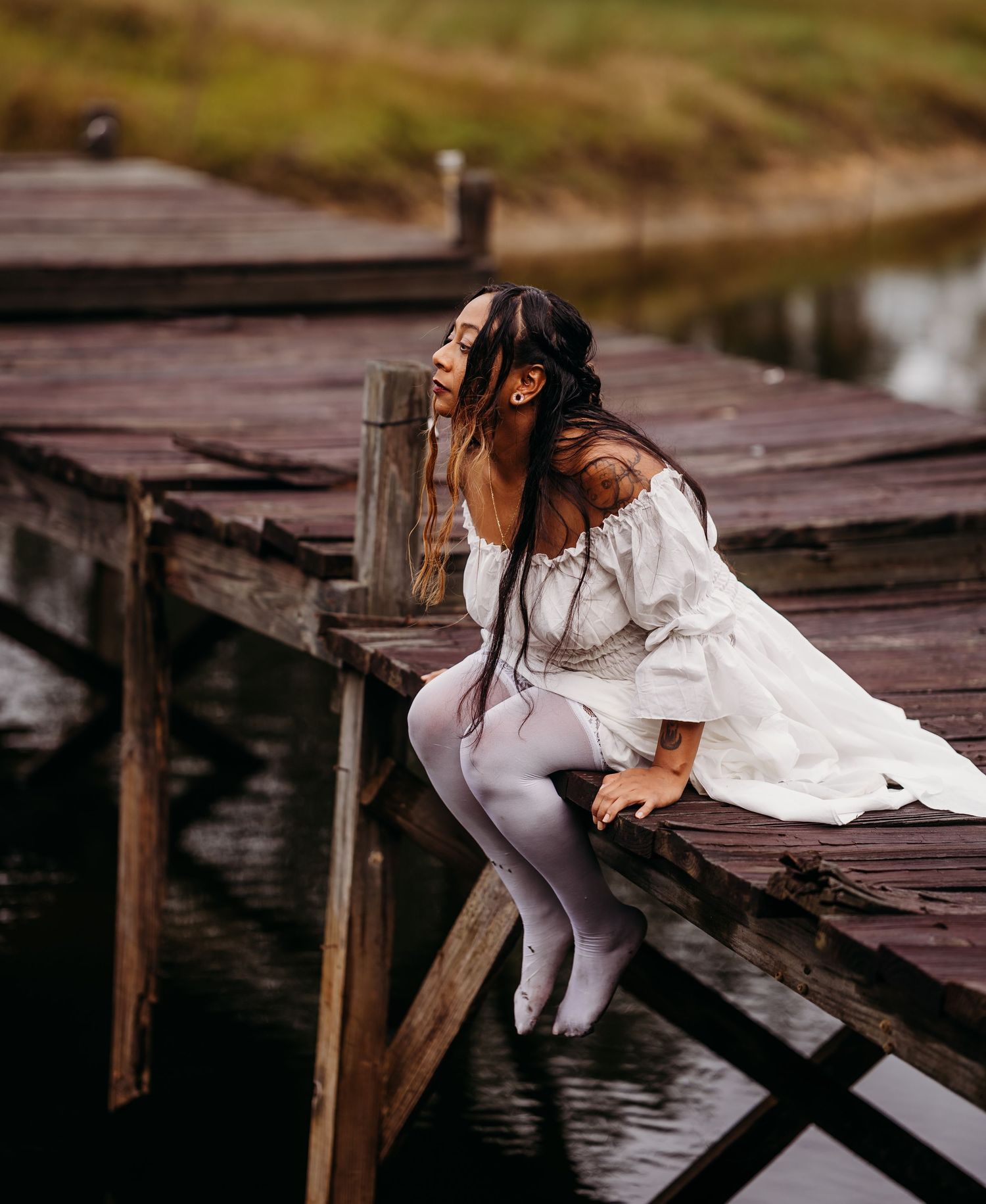 Woman in white off-shoulder dress and white tights sitting on rustic wooden dock over calm water.