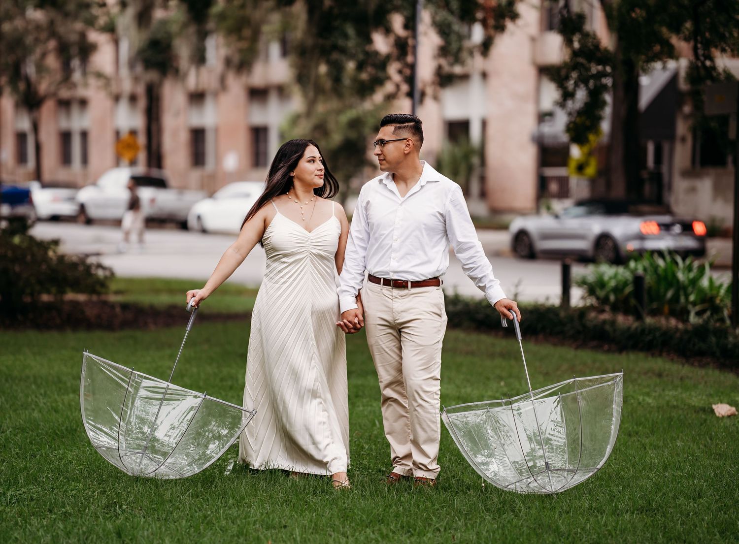 Couple walking hand-in-hand on green grass holding clear bubble umbrellas, woman in white maxi dress, man in white shirt.