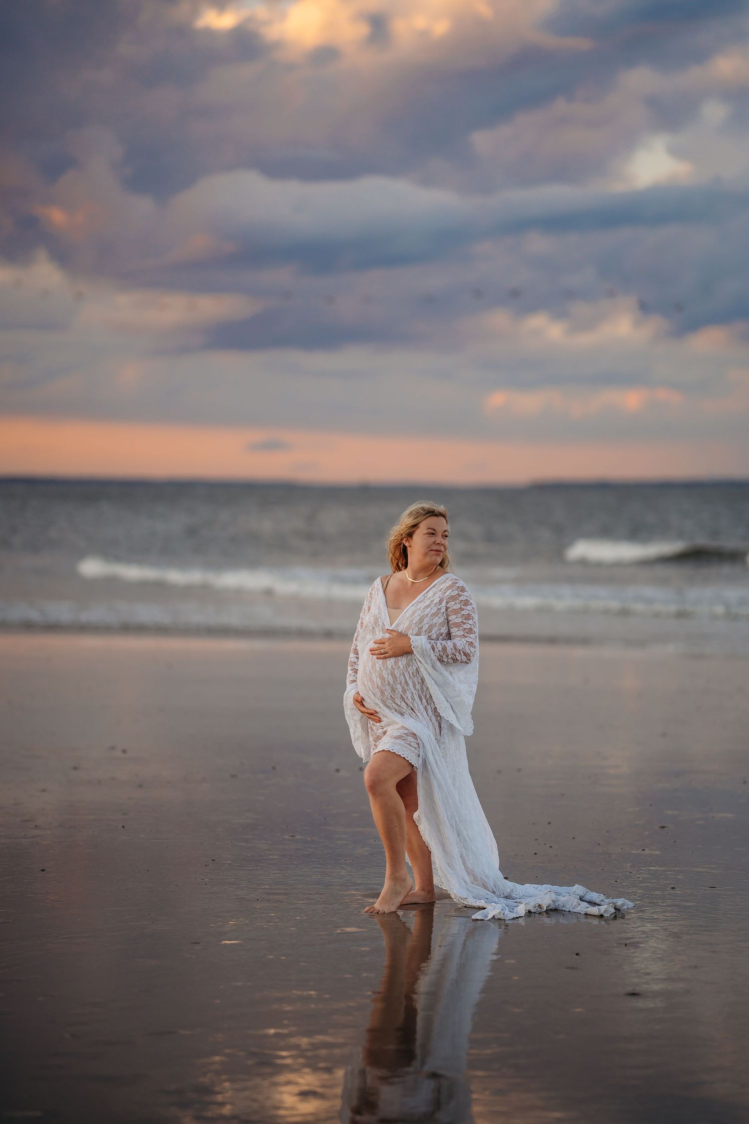 Pregnant woman in white lace gown stands on beach at sunset with waves behind her.