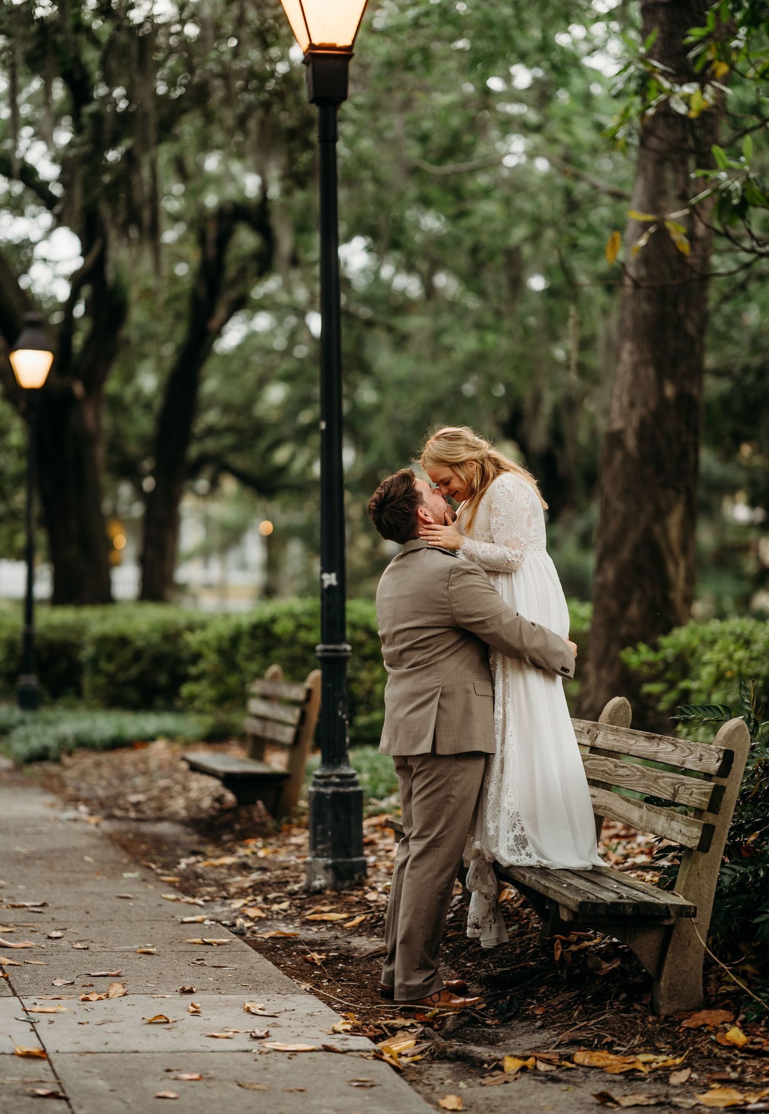 Newlyweds share an intimate kiss beside a glowing lamppost surrounded by autumn leaves and benches.