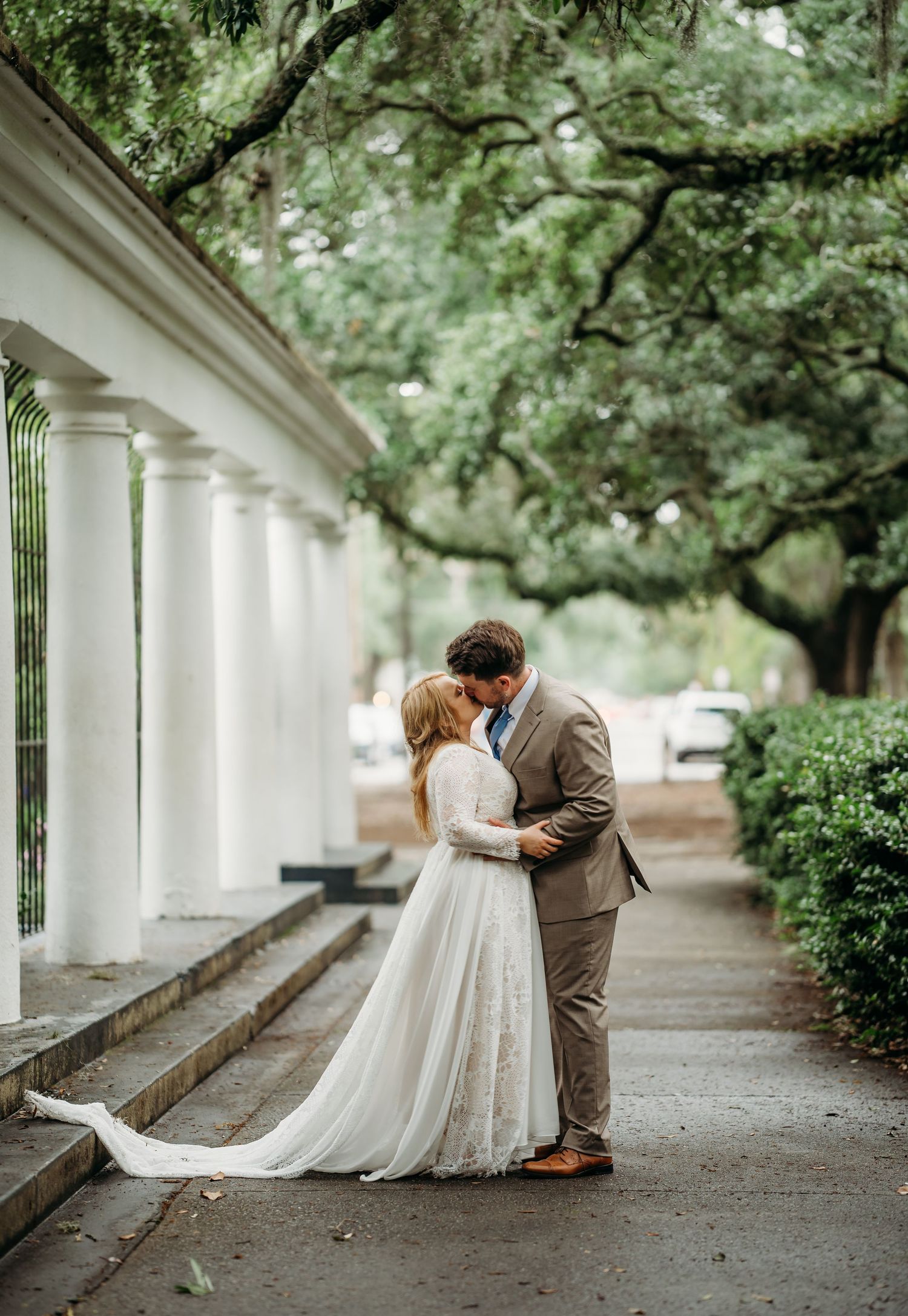 Bride and groom kissing on a charming tree-canopied walkway with white columns and lush green surroundings.