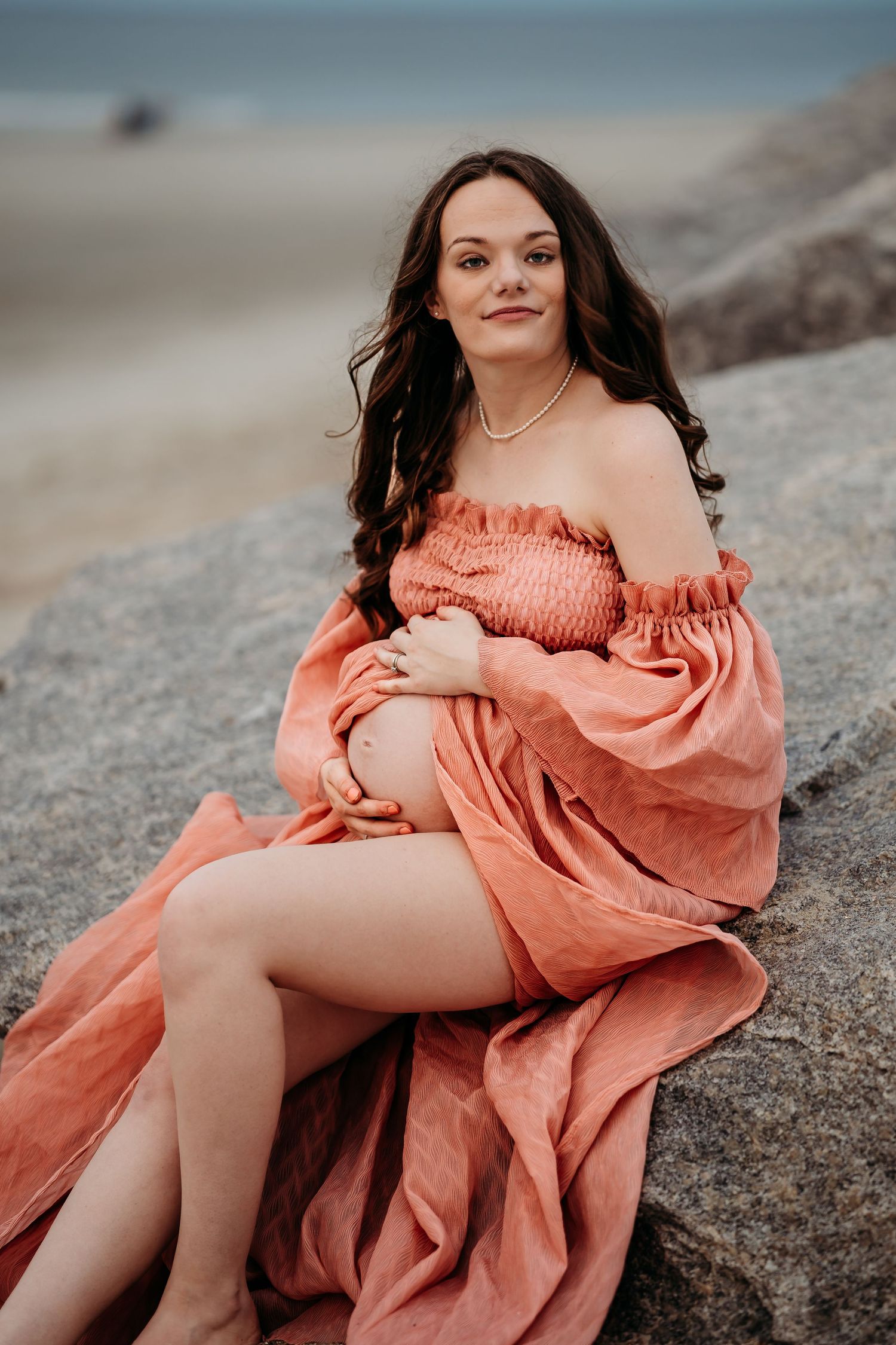 Pregnant woman in off-shoulder coral dress sits on rocks during maternity photoshoot at the beach.