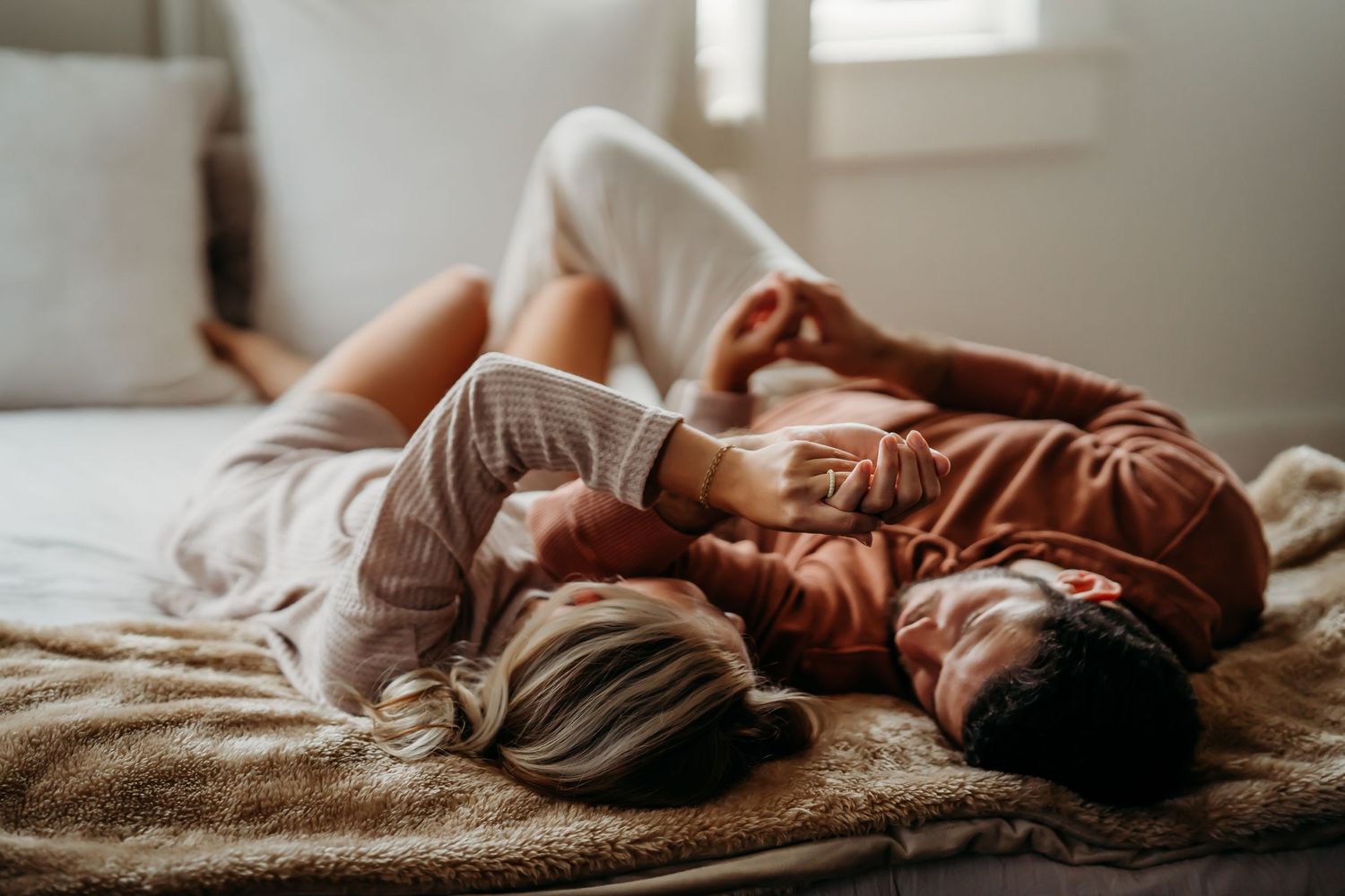 Romantic couple lying side by side on a bed, holding hands in a bright room.