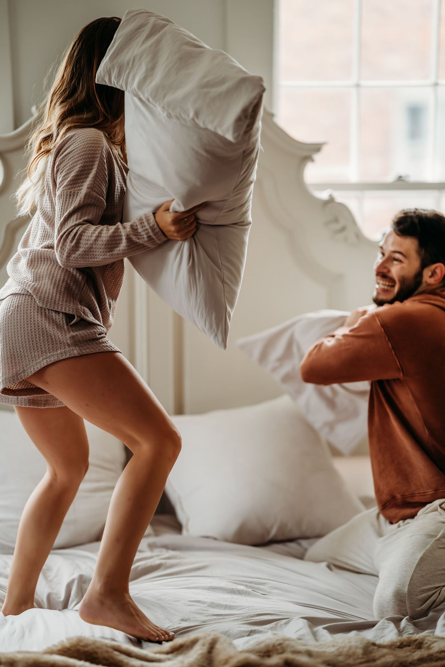 Couple having a playful pillow fight on a white ornate bed, laughing joyfully.