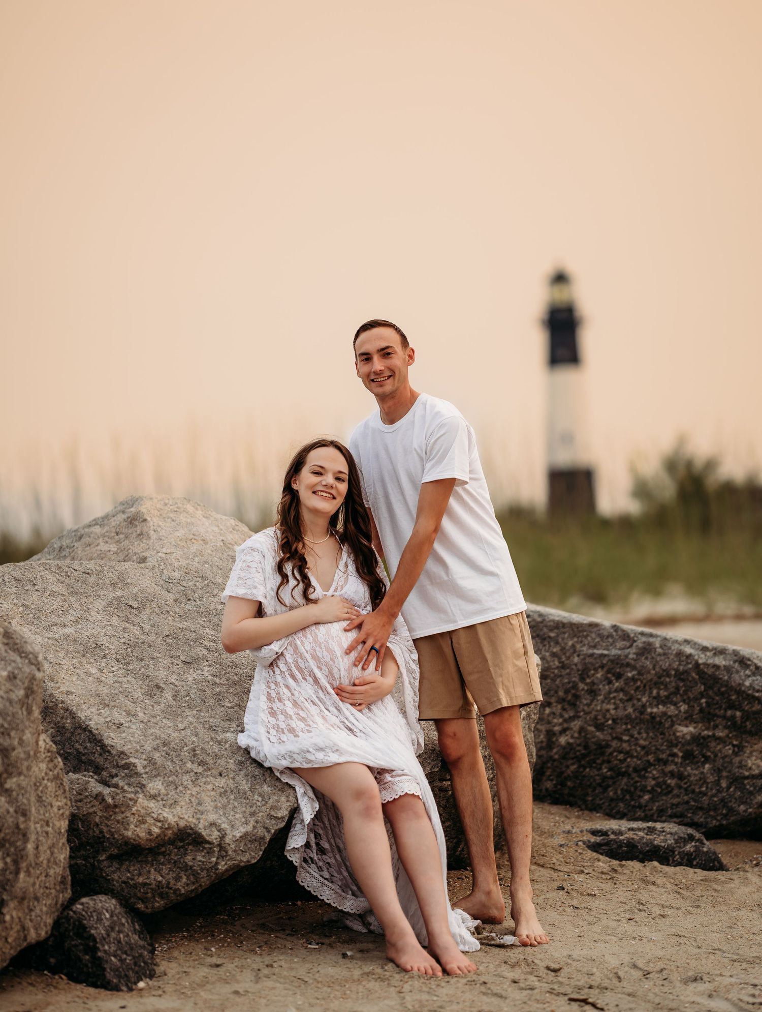 Happy pregnant couple posing on rocky beach at sunset with iconic lighthouse backdrop, woman in white lace dress.