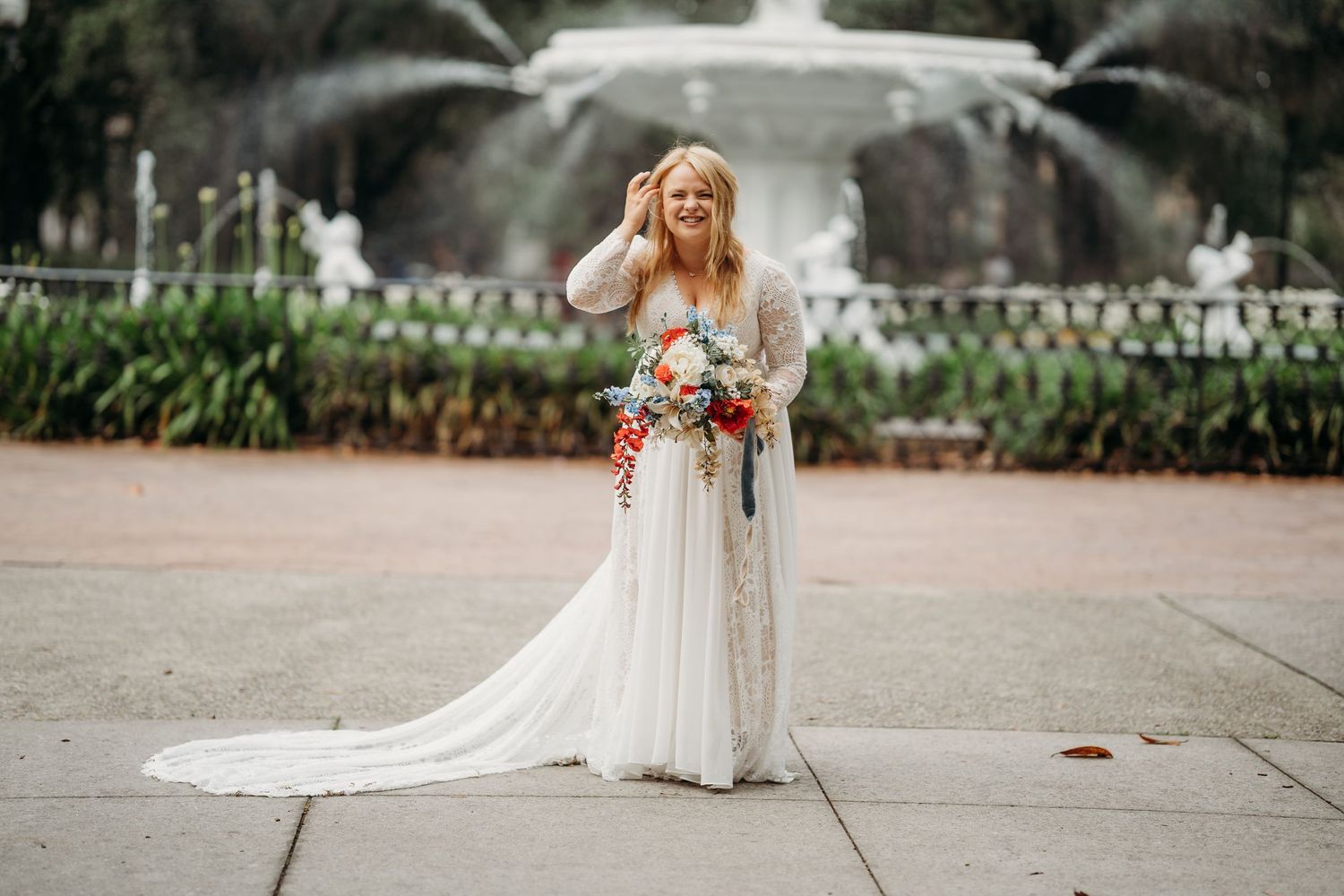 Blonde bride in white lace gown laughs joyfully holding colorful bouquet before a park fountain.