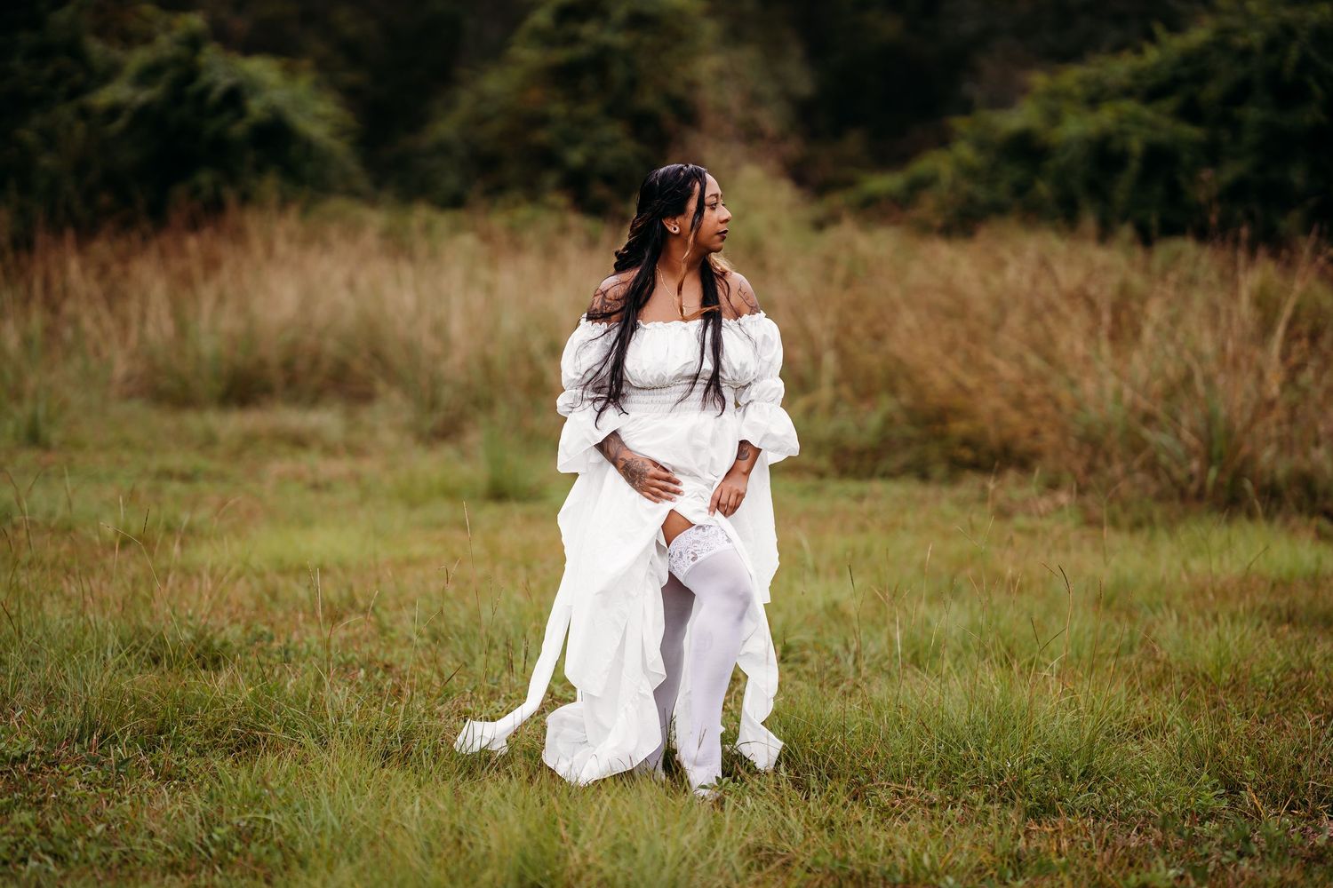 Woman in white layered dress sitting elegantly in grassy field with forest background.