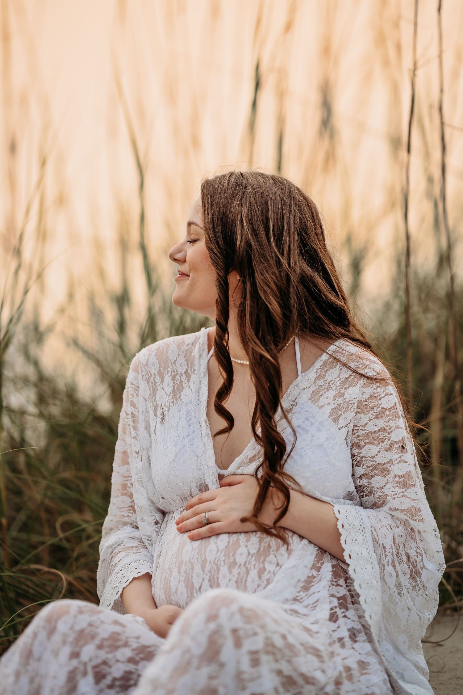 Pregnant woman in white lace dress sitting on beach sand smiling at golden sunset.