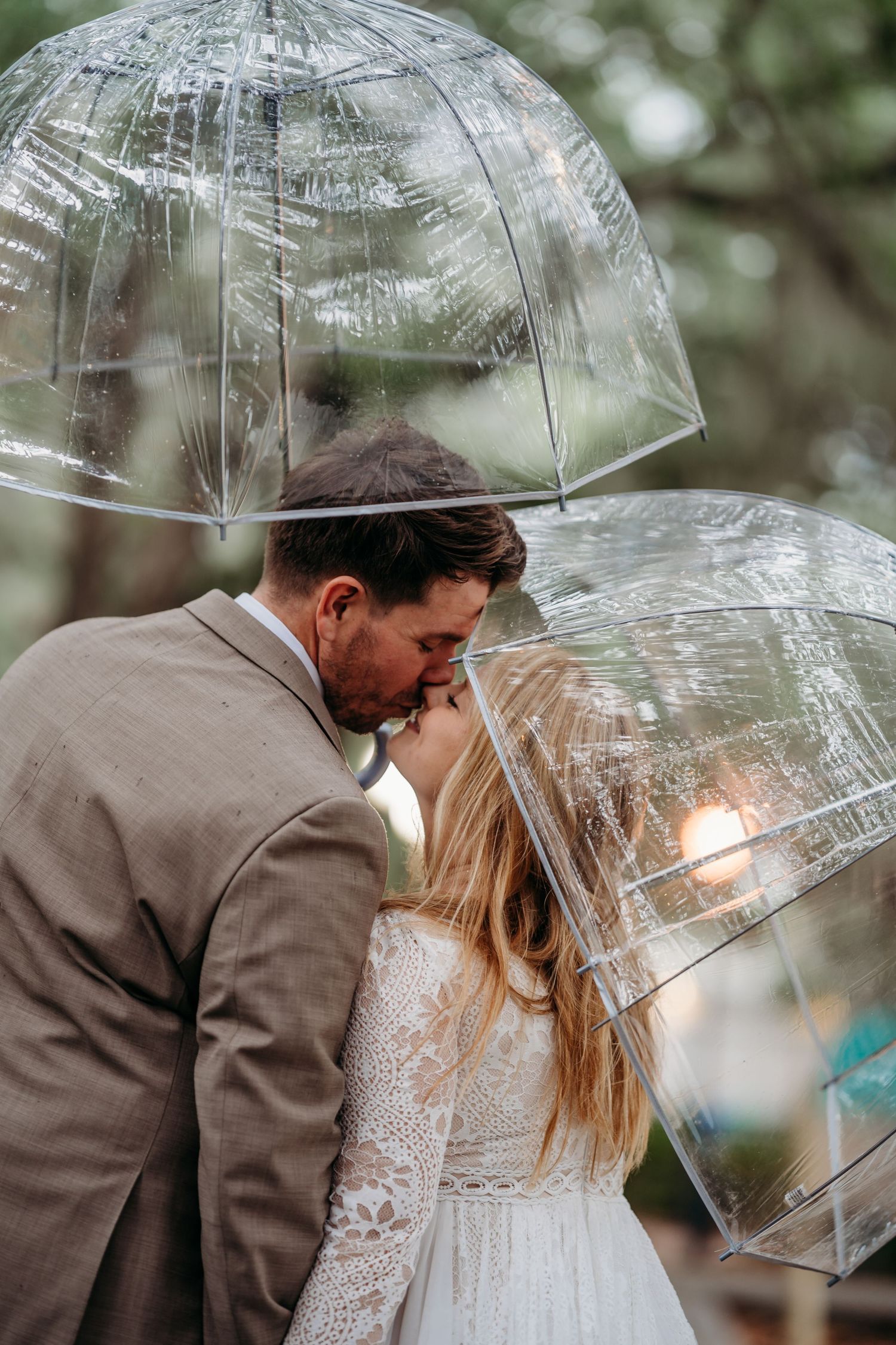 Bride and groom sharing a kiss under clear bubble umbrellas in a lush outdoor setting on a rainy wedding day.