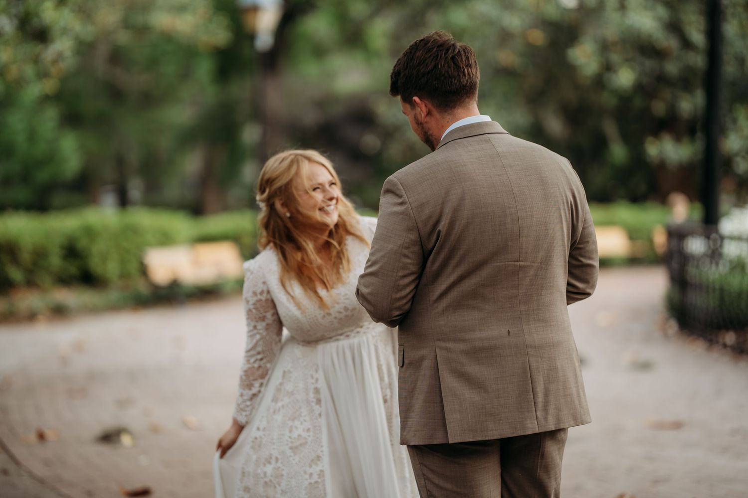Groom in tan suit gently touches bride's shoulder during romantic outdoor wedding in lush park.