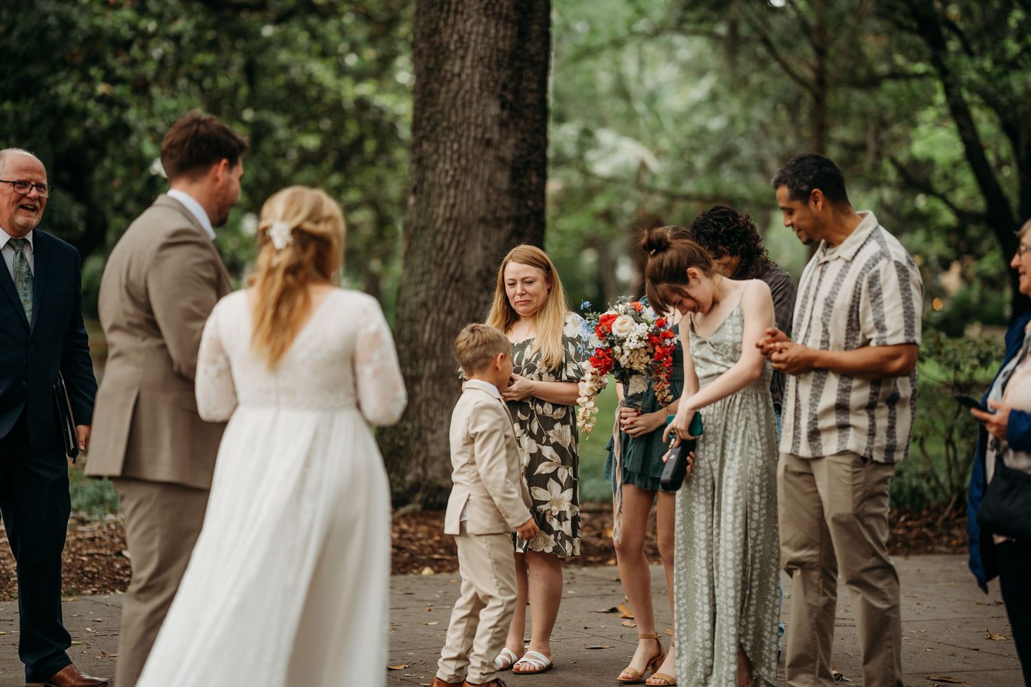 Bride and groom greet family members including a young child holding red flowers in a park ceremony.