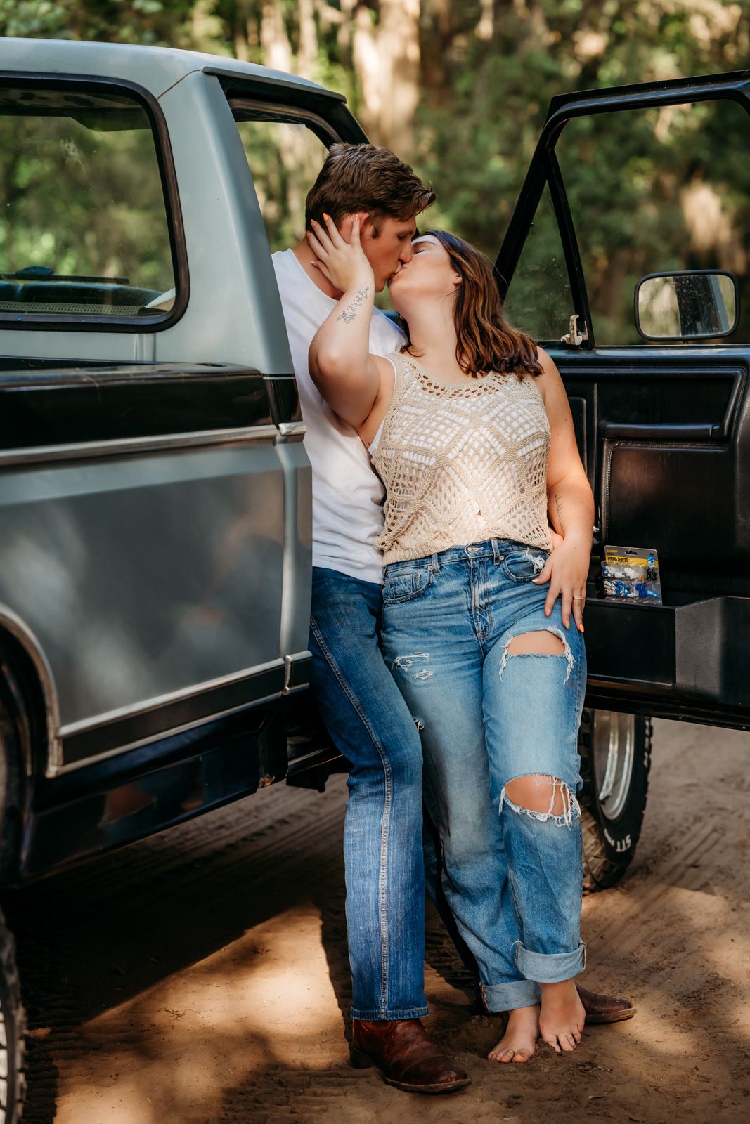 Couple shares romantic kiss leaning against vintage black pickup truck in wooded setting.