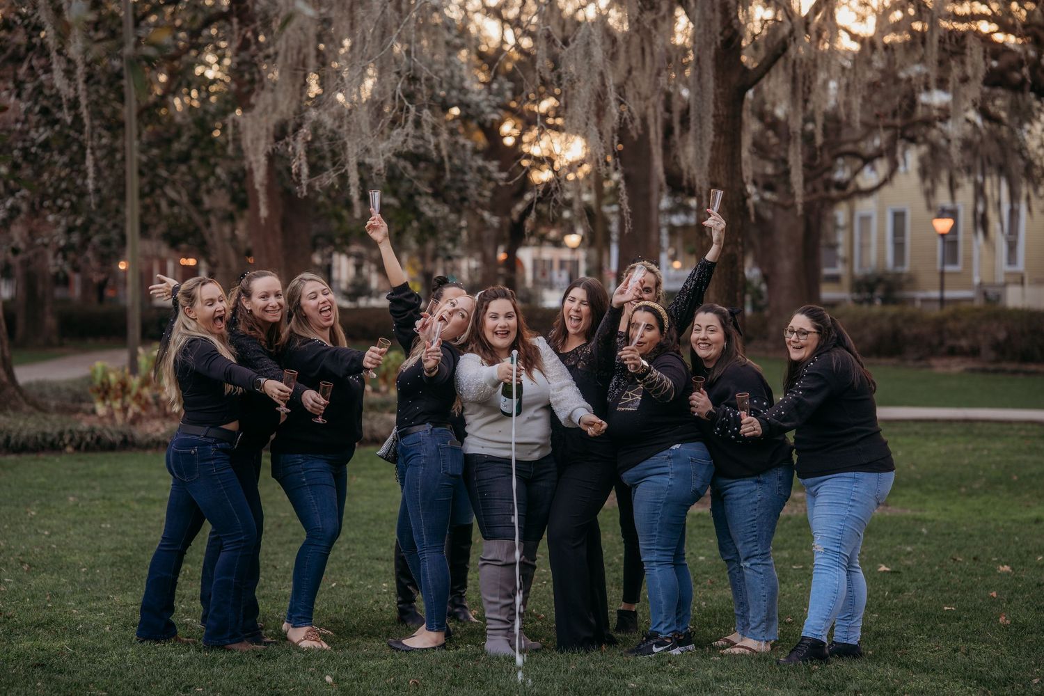 Group of women posing and laughing outdoors under Spanish moss trees at sunset in a park.