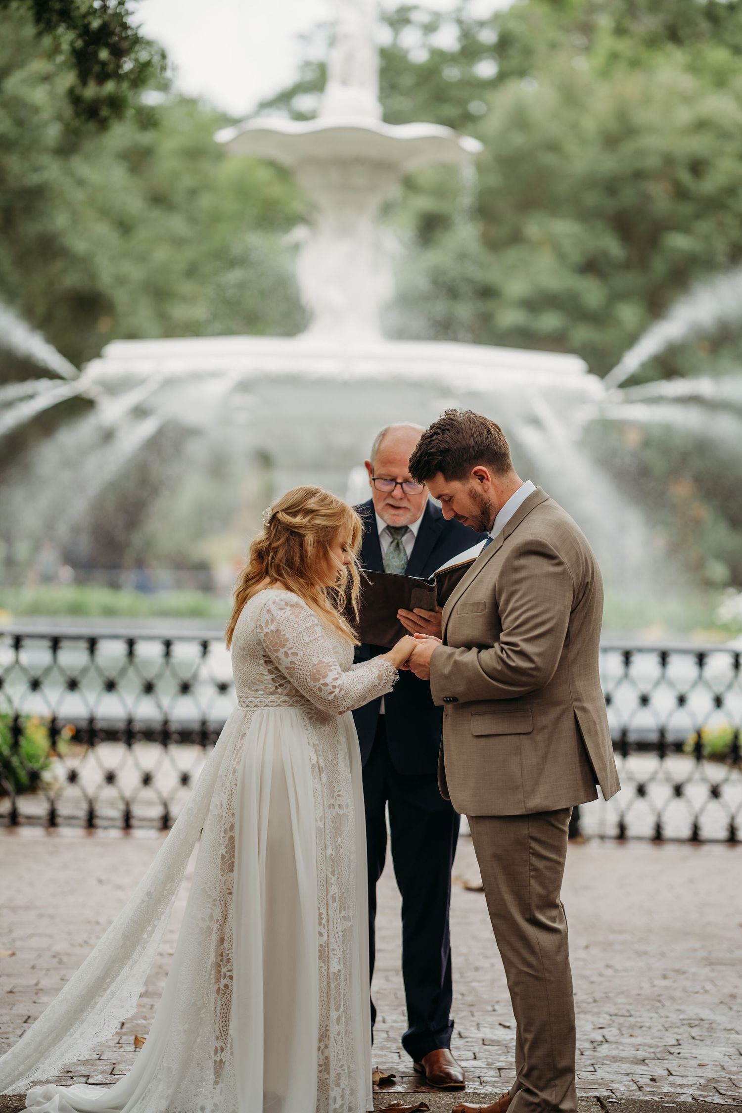 Groom places wedding ring on brides finger before ornate white fountain during outdoor park wedding ceremony.