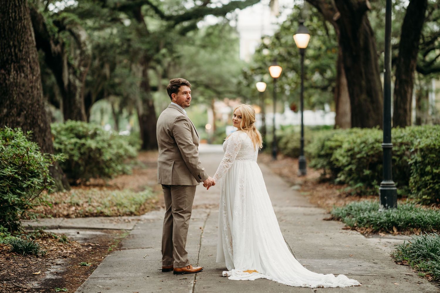 Bride in long-sleeve white gown and groom in tan suit hold hands facing each other on a glowing lantern-lit path.