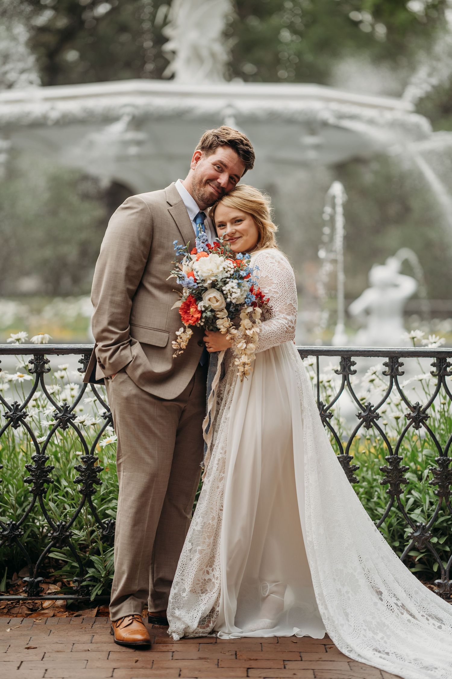 Bride leans on groom holding a vibrant bouquet while posing before an ornate fountain backdrop.