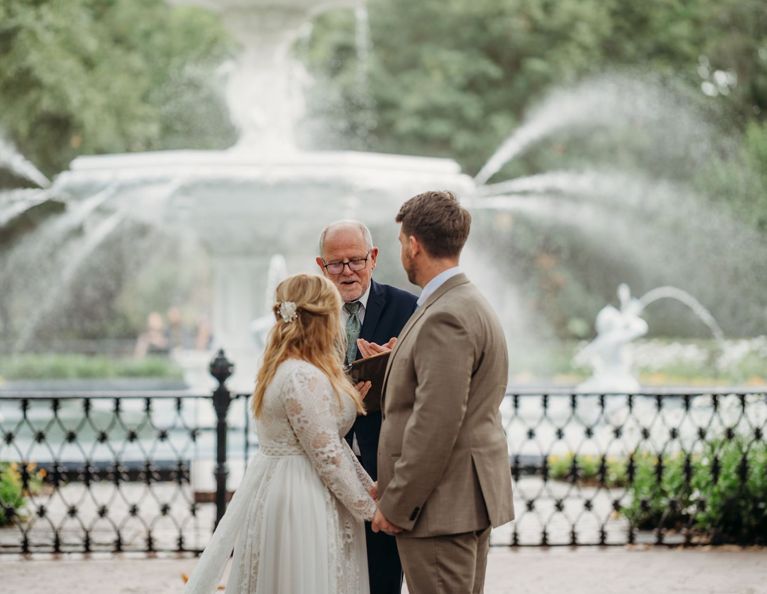 Couple stands with officiant during outdoor wedding ceremony in front of ornate park fountain.