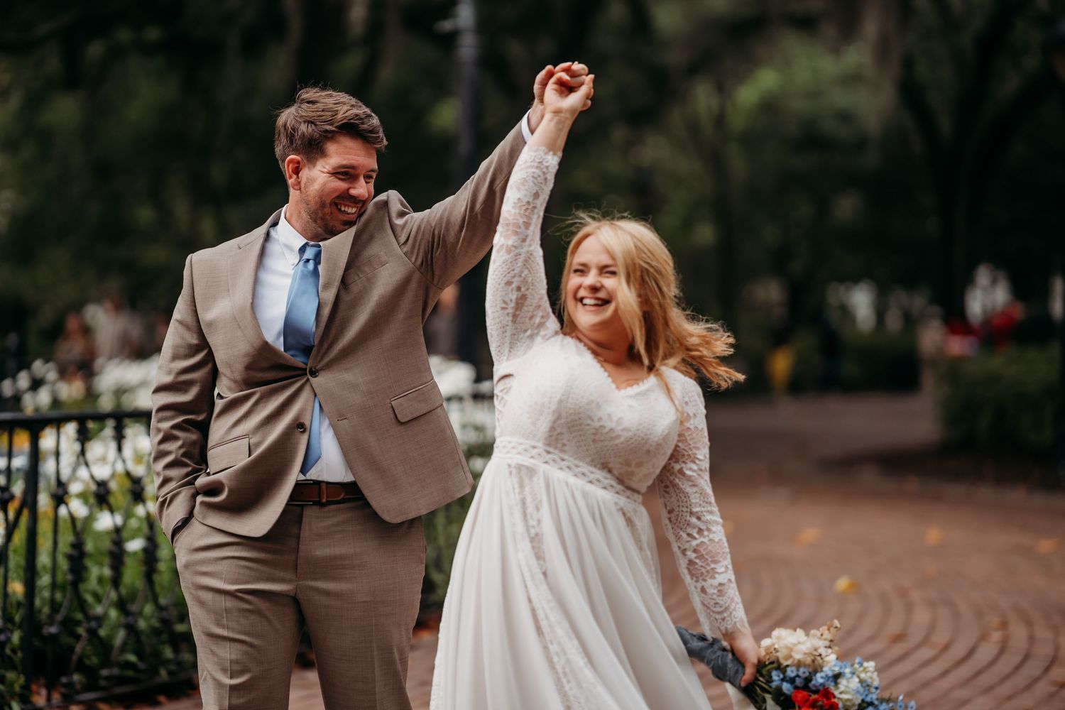 Groom twirls bride in a white lace gown on a brick path, both smiling joyfully with colorful bouquet in hand.