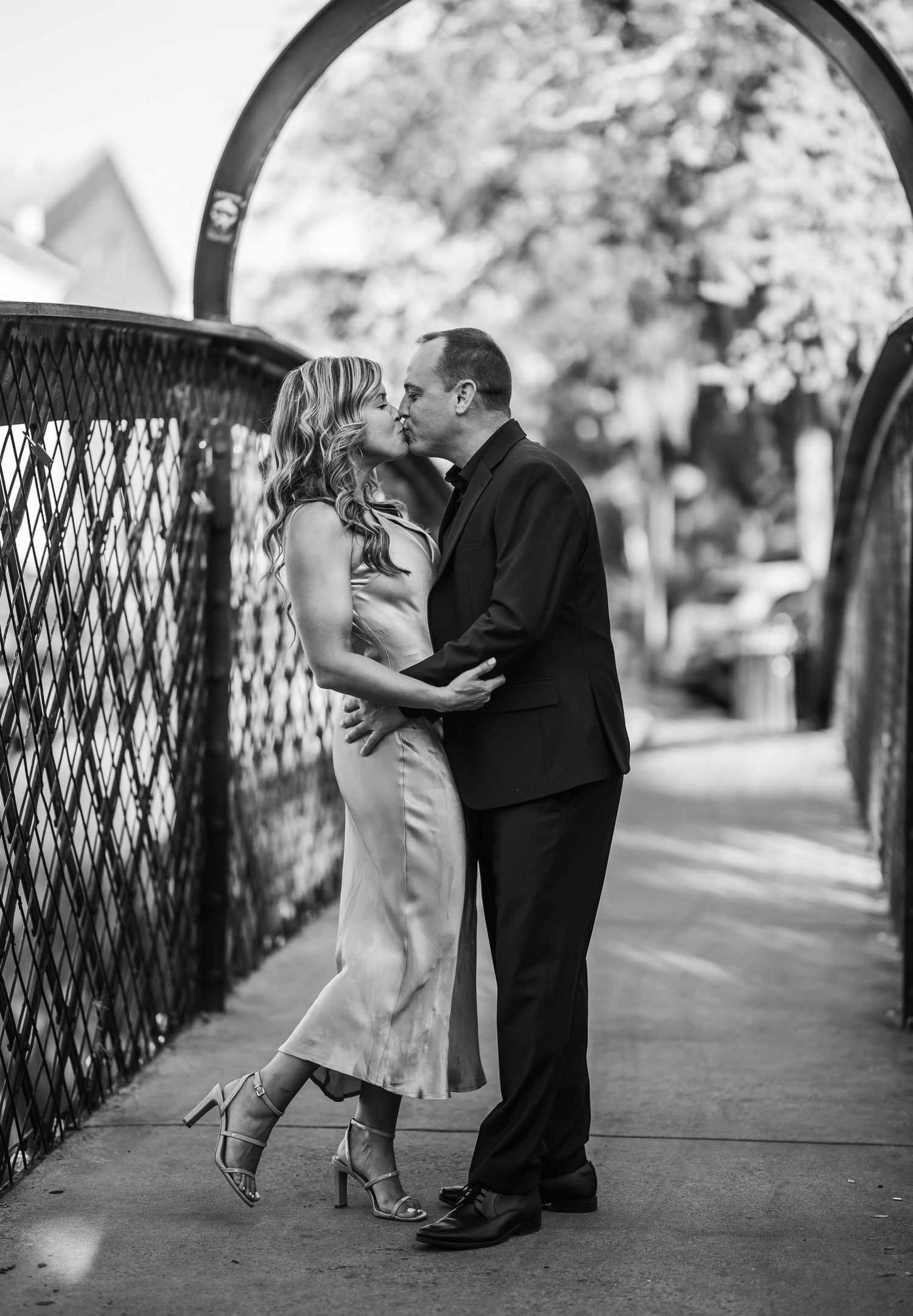Black and white photo of a couple sharing a romantic kiss on a bridge walkway, woman in midi dress and heels.