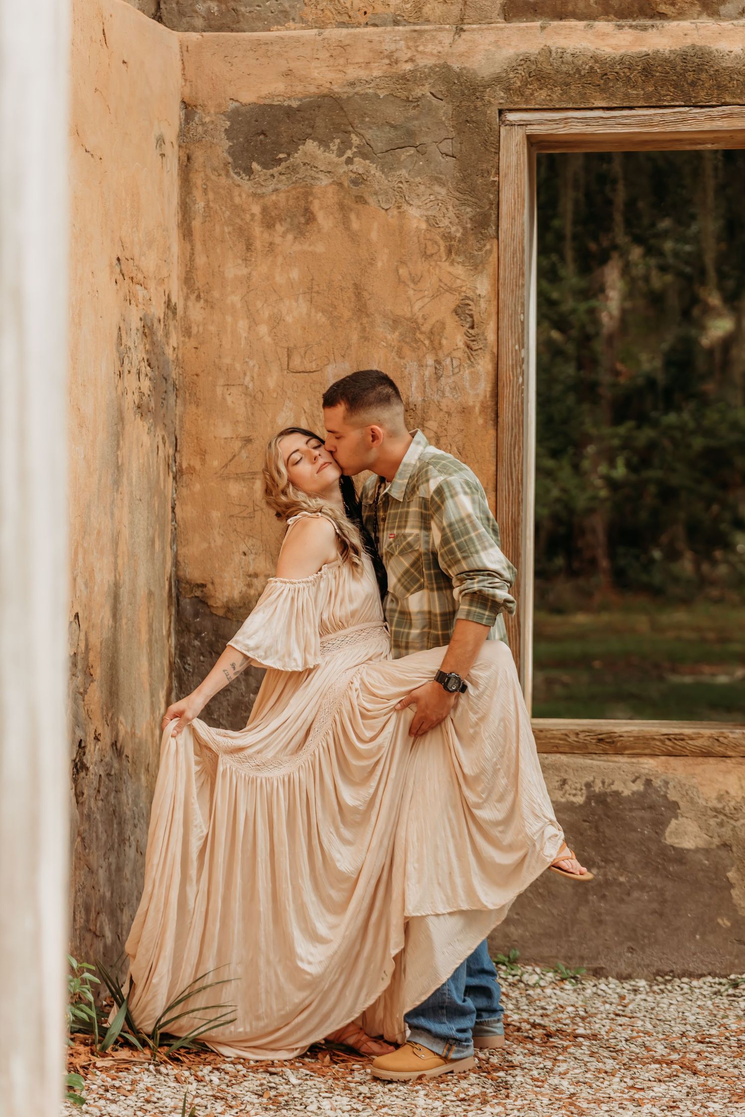 Couple sharing a romantic kiss against a rustic weathered wall, woman in flowing blush gown, man in plaid shirt and jeans.