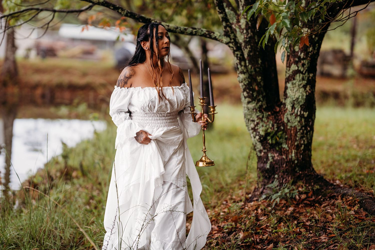 Woman in white off-shoulder gown holds gold candelabra beside mossy tree near pond in autumn park.