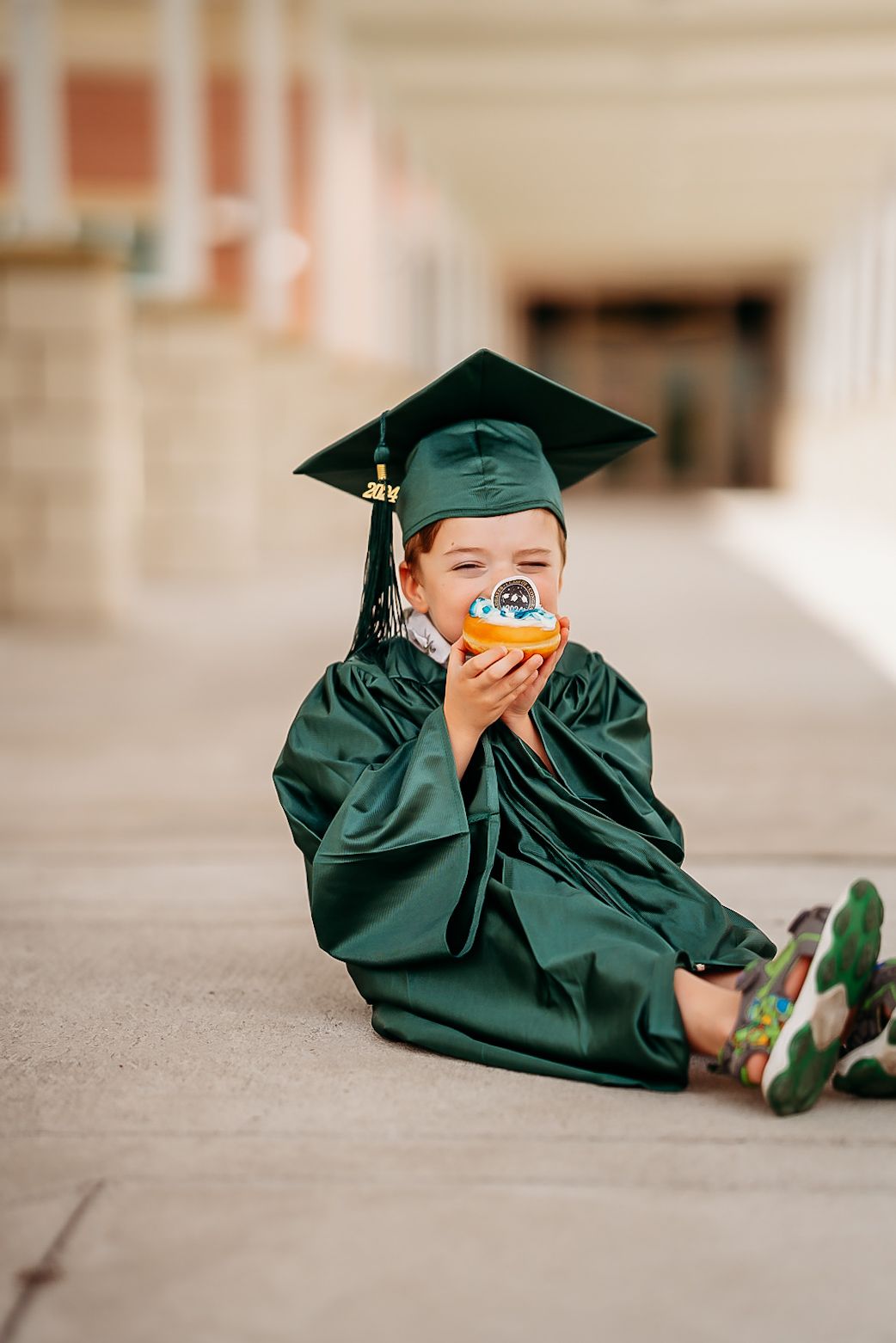 Adorable child in green graduation cap and gown eating a colorful donut while sitting outdoors.