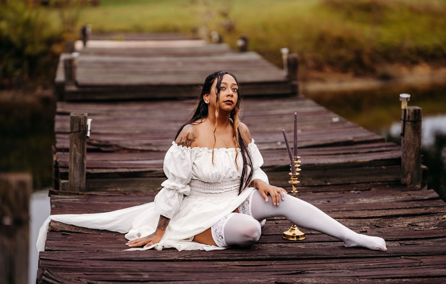 Woman in white dress and white stockings sits in splits on wooden pier with candle nearby.
