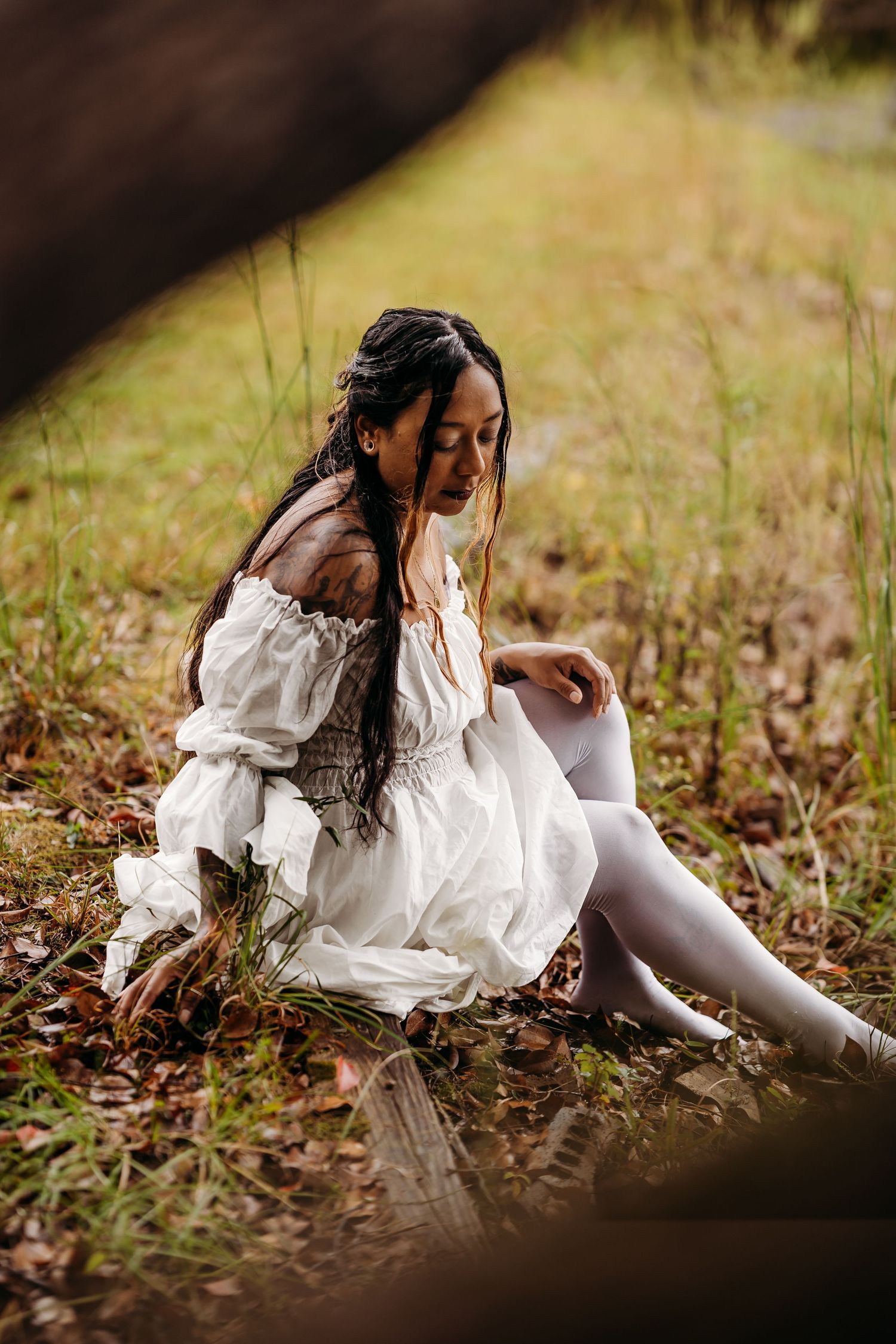 Woman in white off-shoulder dress seated on ground surrounded by autumn leaves outdoors.
