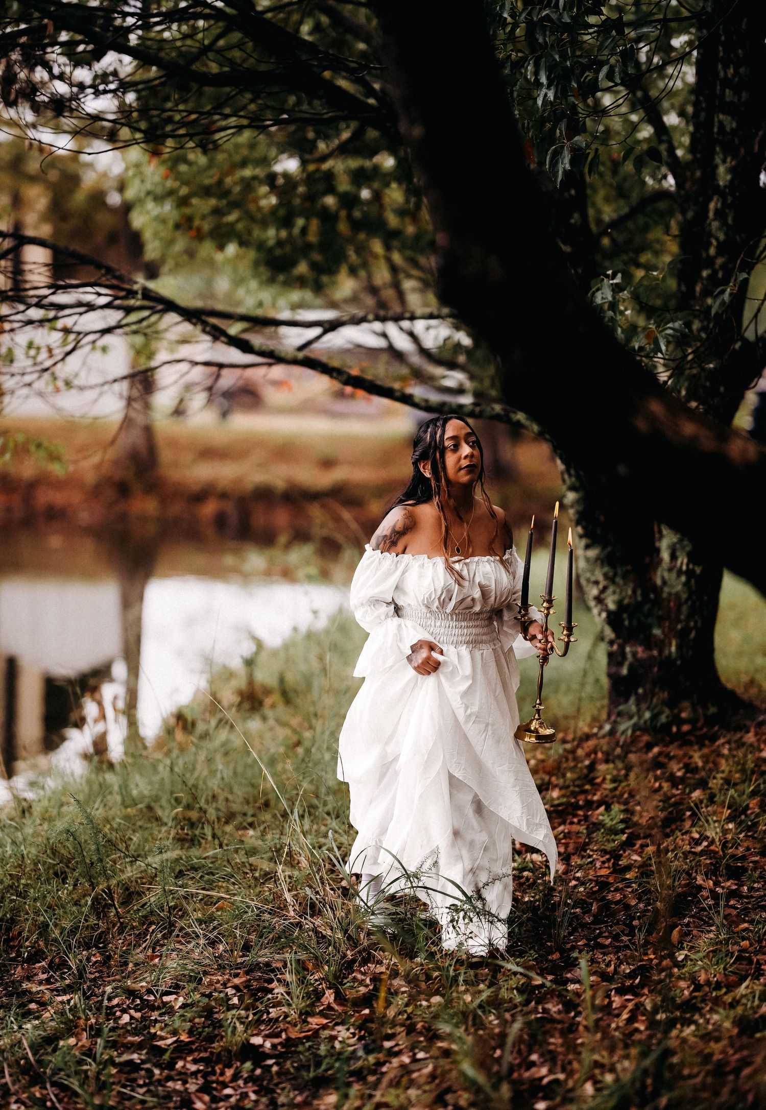 Woman in white off-shoulder dress holding candelabra by autumn pond.