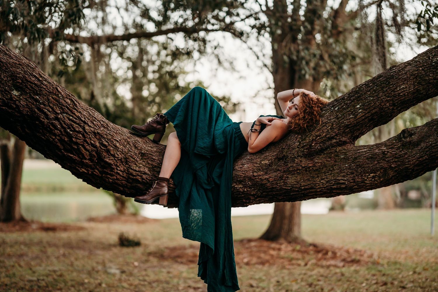 Woman in emerald green dress posing dramatically draped across a large curved tree branch outdoors.
