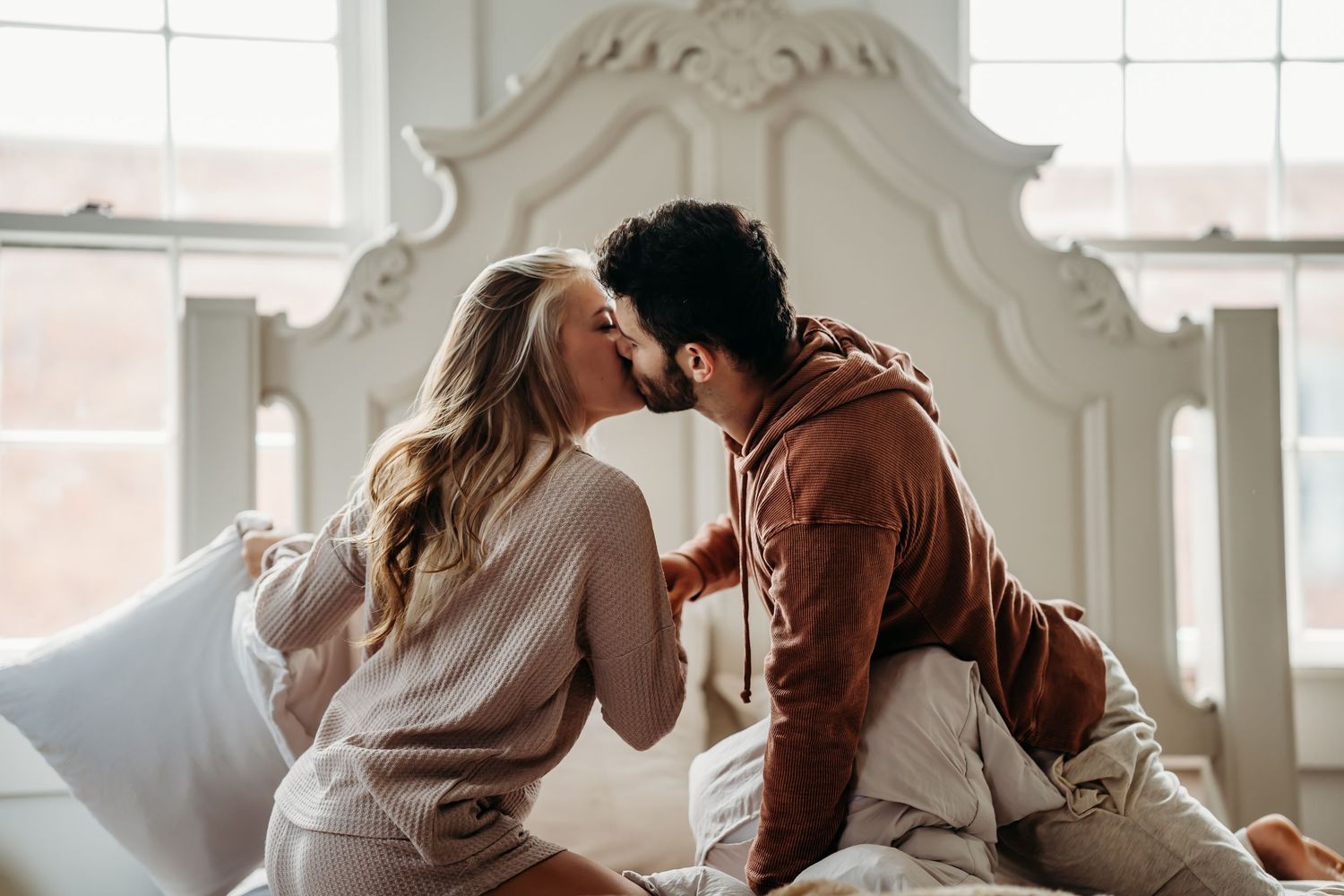 A couple sharing a tender kiss while sitting on a white bed near a sunlit window.