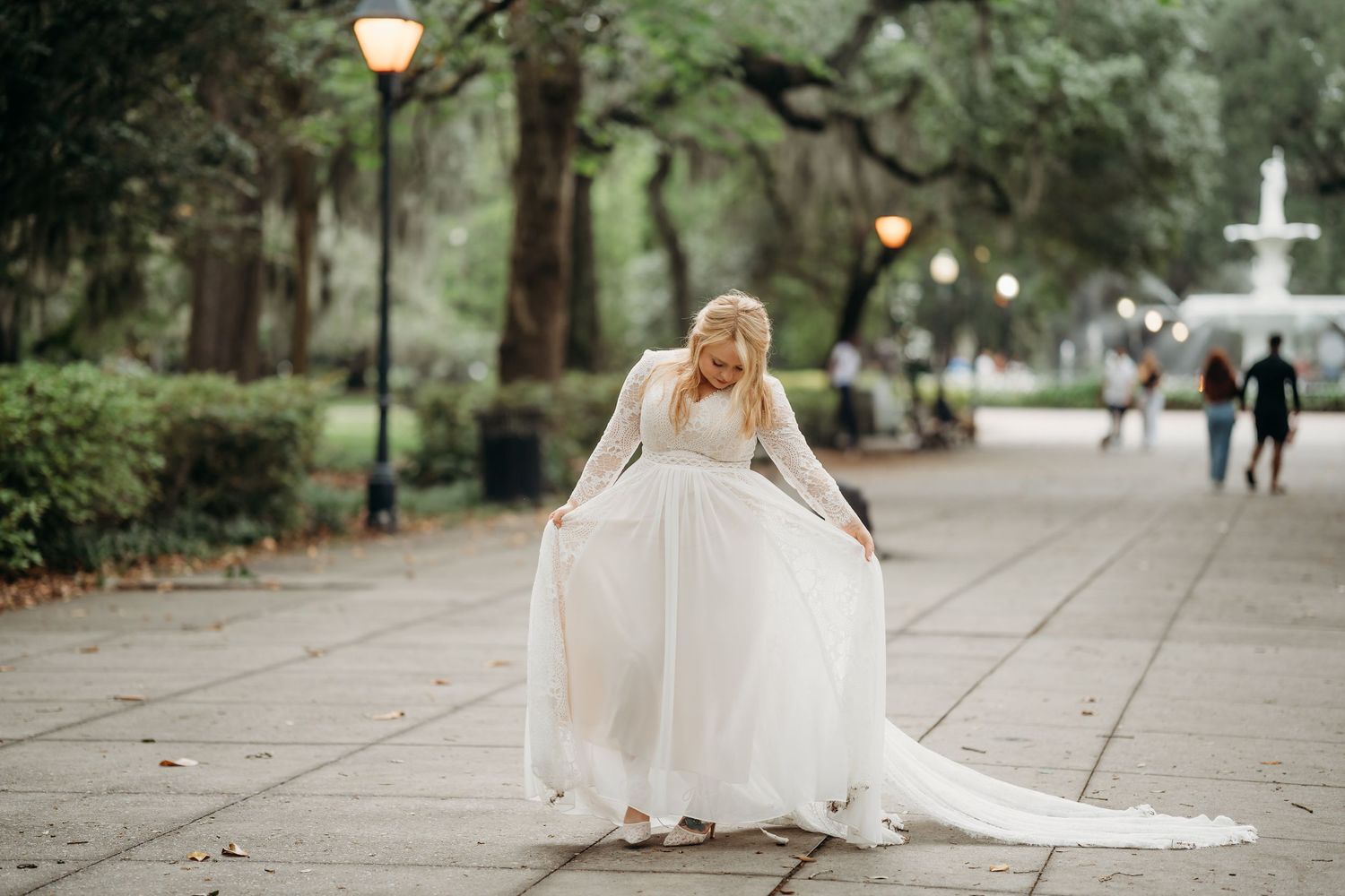 Bride in lace long-sleeve gown twirls on Forsyth Park pathway near fountain in Savannah, Georgia.