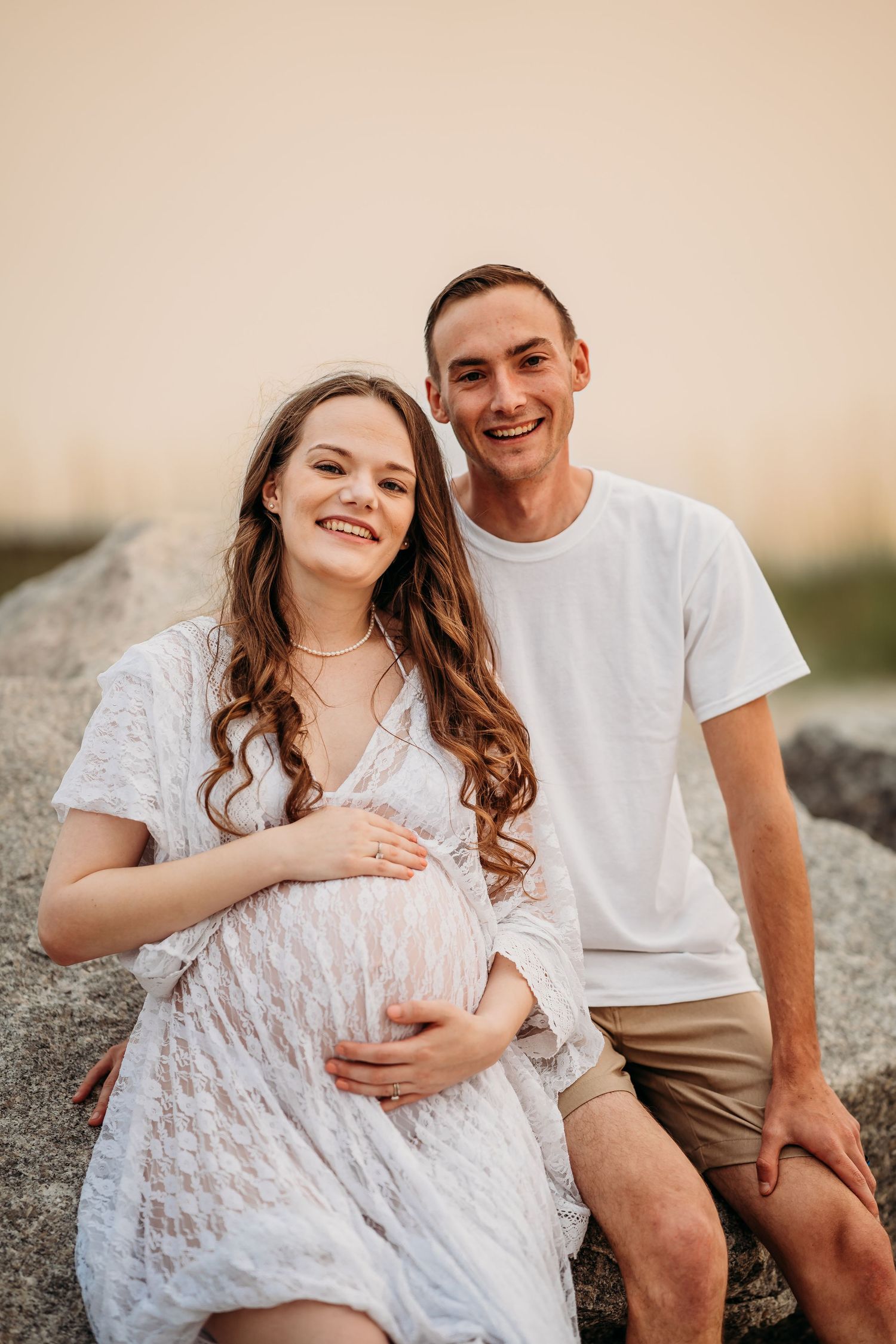 Smiling pregnant couple sitting on rocks at sunset, woman in white lace maternity dress cradling baby bump.